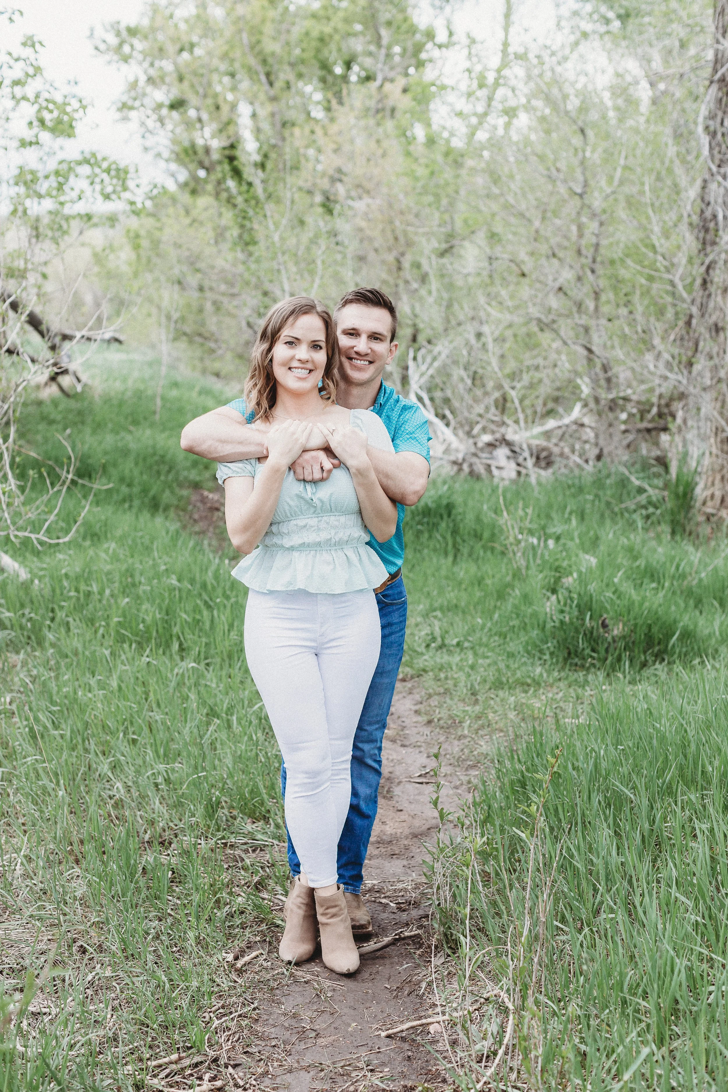 A smiling couple standing on a narrow dirt path through a green wooded area during daytime, with the woman in a light blue top, white pants, and beige ankle boots, and the man in a blue shirt and jeans, embracing behind her.