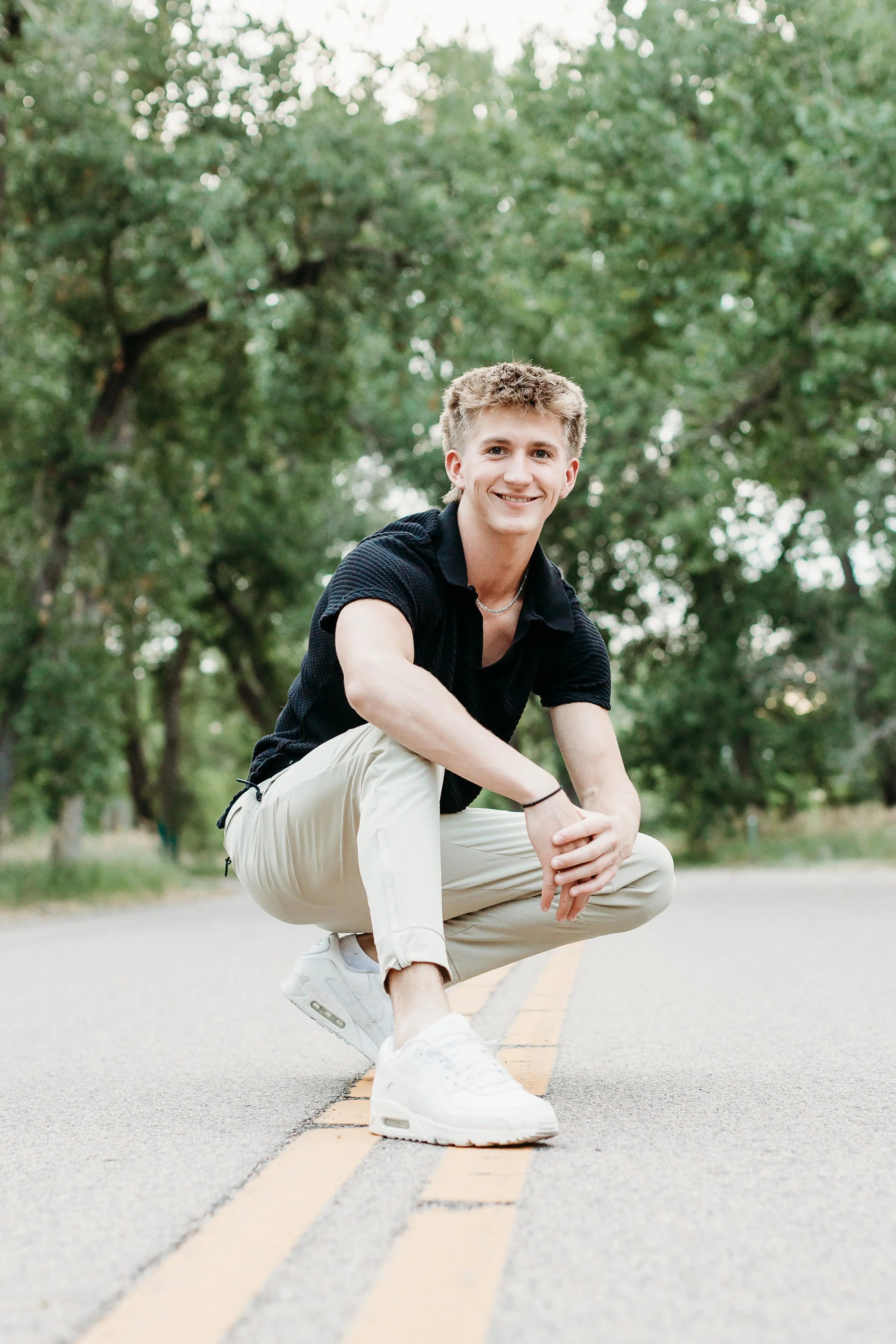A young man with short blond hair squatting on a road with double yellow lines, smiling at the camera, surrounded by green trees.