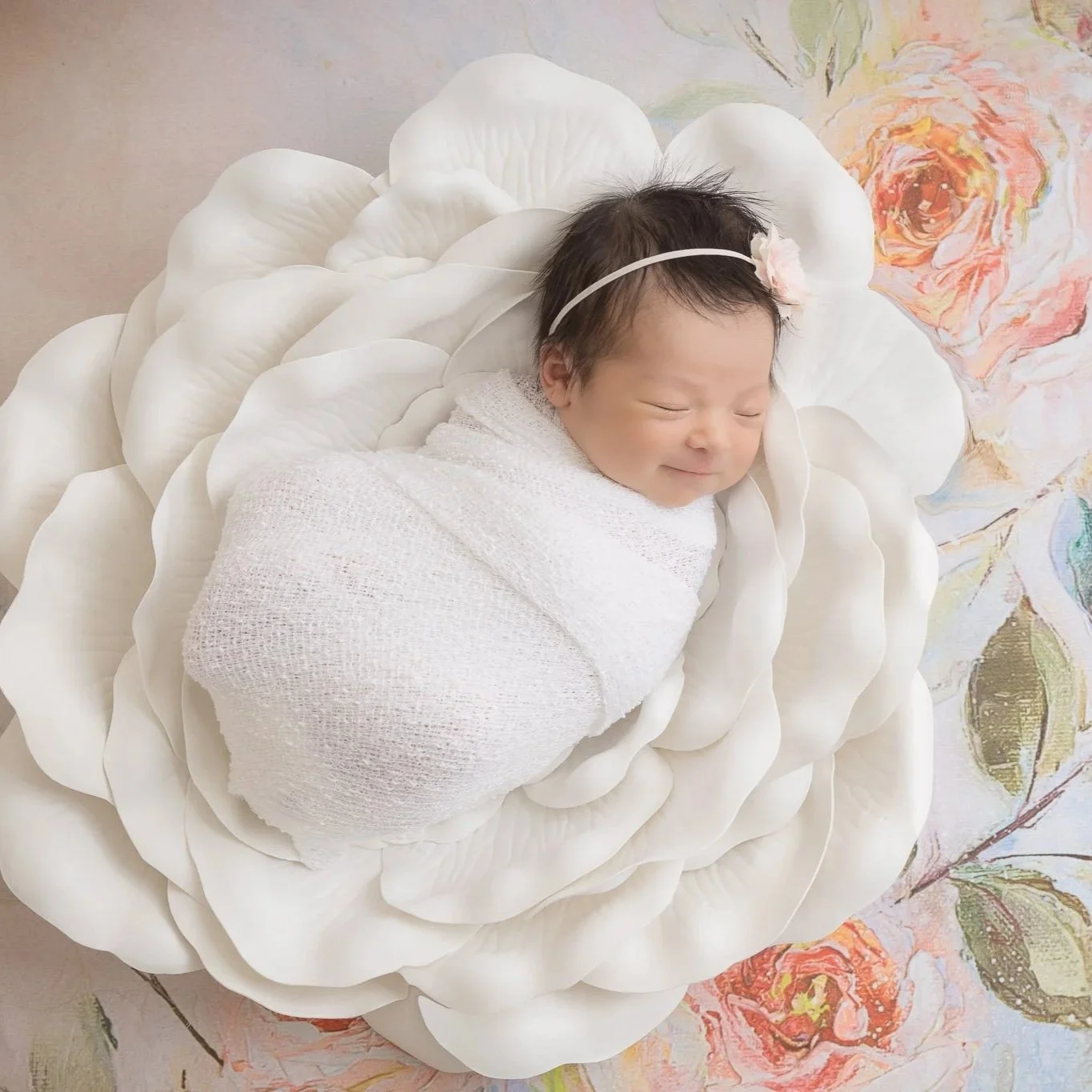A smiling baby swaddled in a white cloth, lying on a large, white, flower-shaped cushion with a pink flower headband on her head, resting on a floral-patterned background.