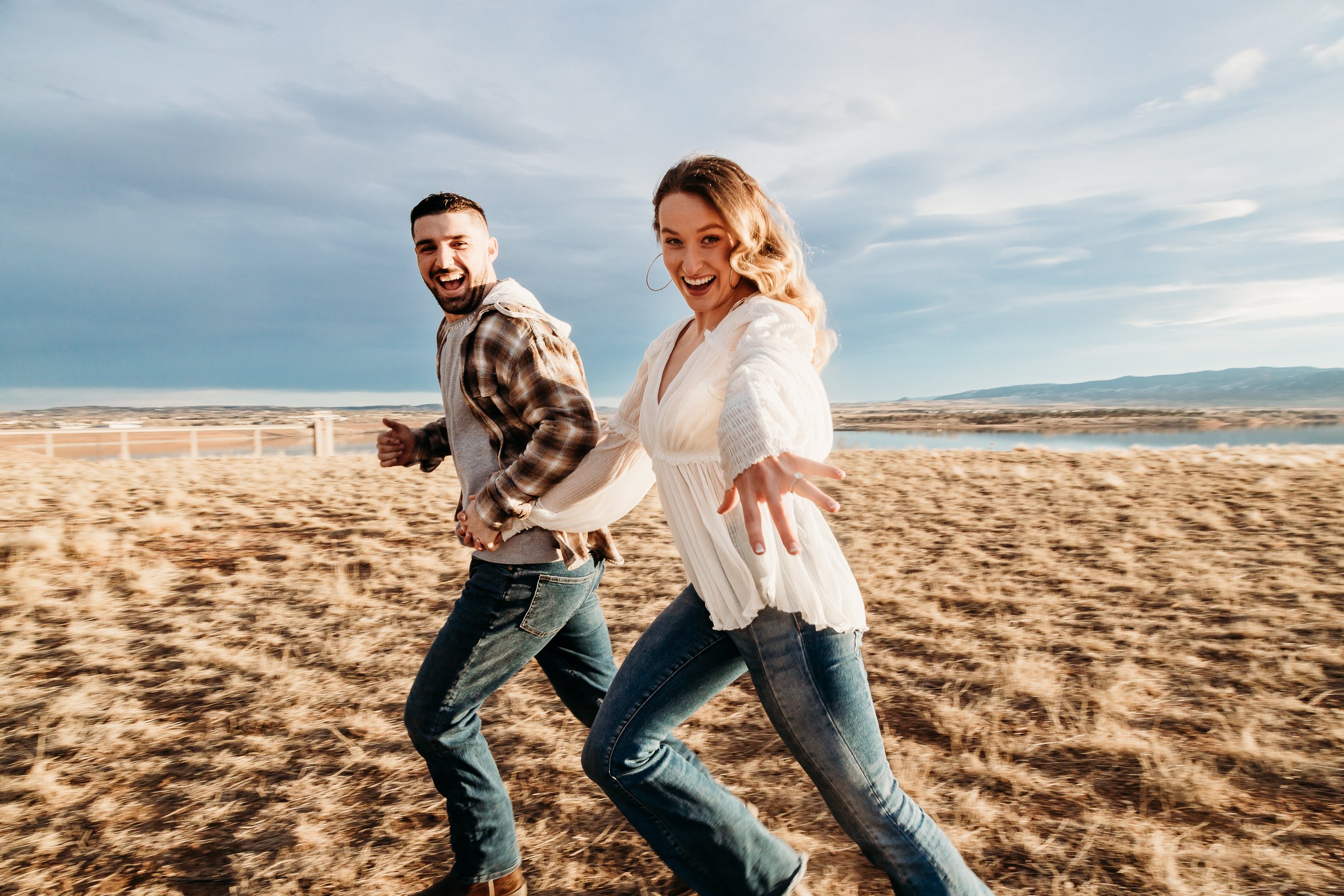 A happy couple running along a sandy beach with a body of water and a cloudy sky in the background.