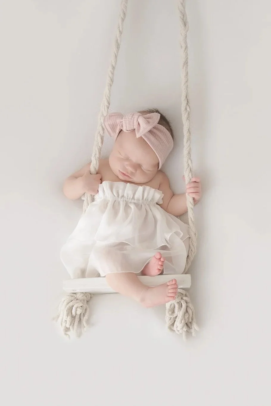 A sleeping baby girl wearing a pink headband with a bow, lying on a white swing with fluffy ropes, dressed in a white dress with ruffle details.