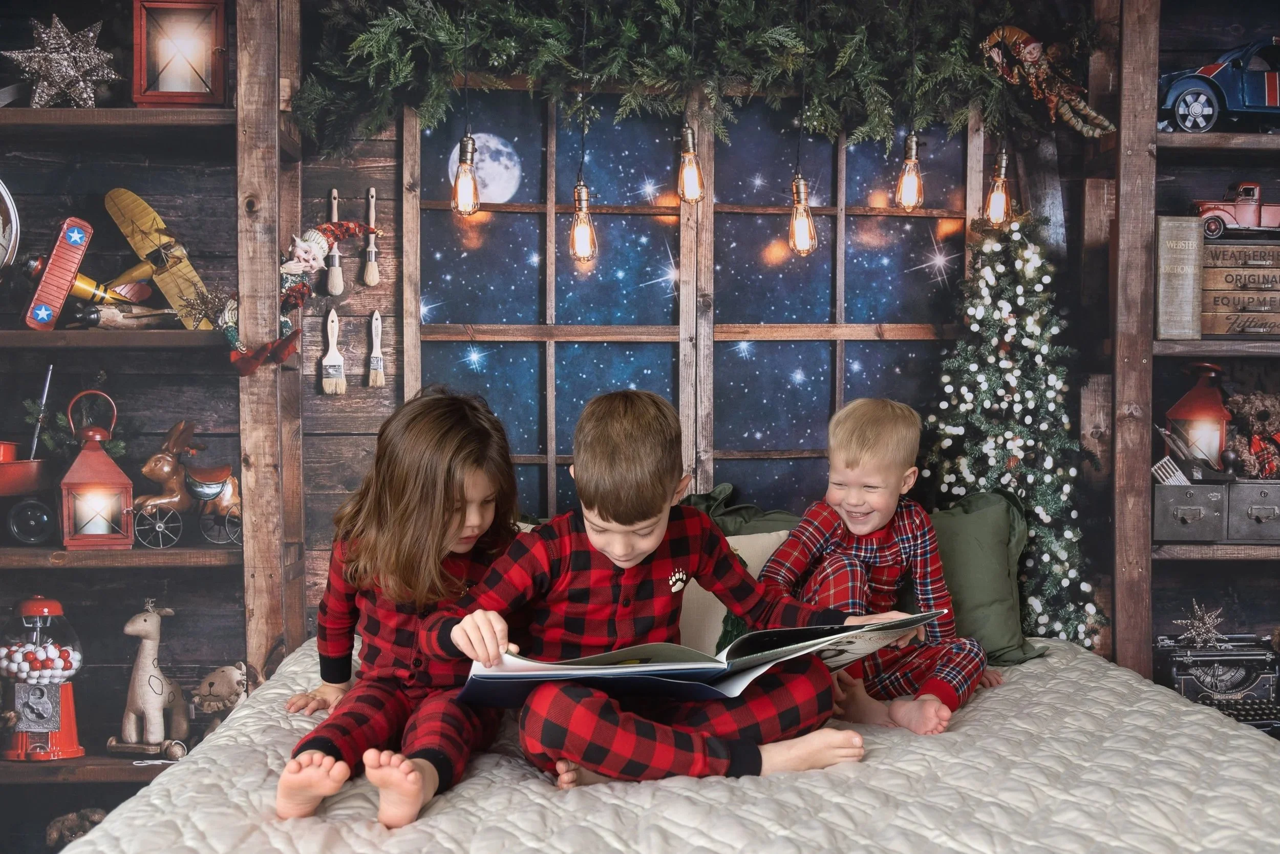 Three children in Christmas pajamas sitting on a bed, reading a book, with a Christmas tree and holiday decorations in the background.