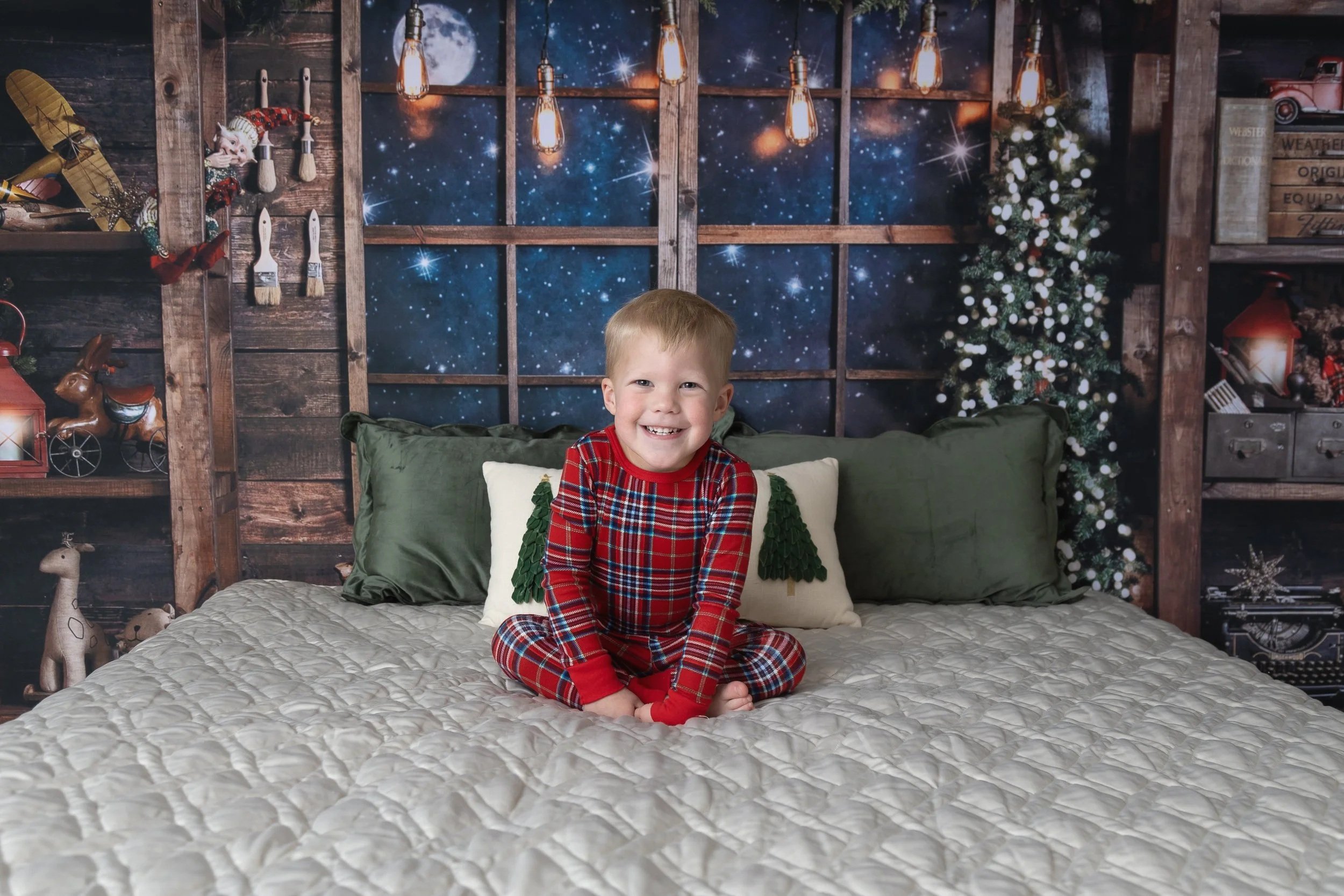 A young boy in red plaid pajamas sitting on a bed with a festive holiday backdrop featuring a starry night sky, Christmas tree, and holiday decorations.
