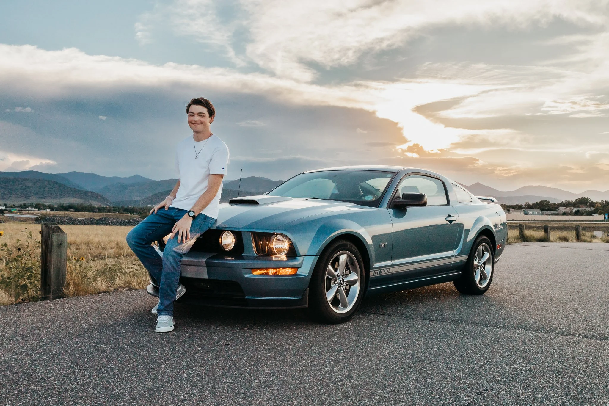 A young man in a white t-shirt and jeans sitting on the hood of a blue Ford Mustang GT 300 parked on a paved road at sunset. Mountains in the background with a partly cloudy sky.