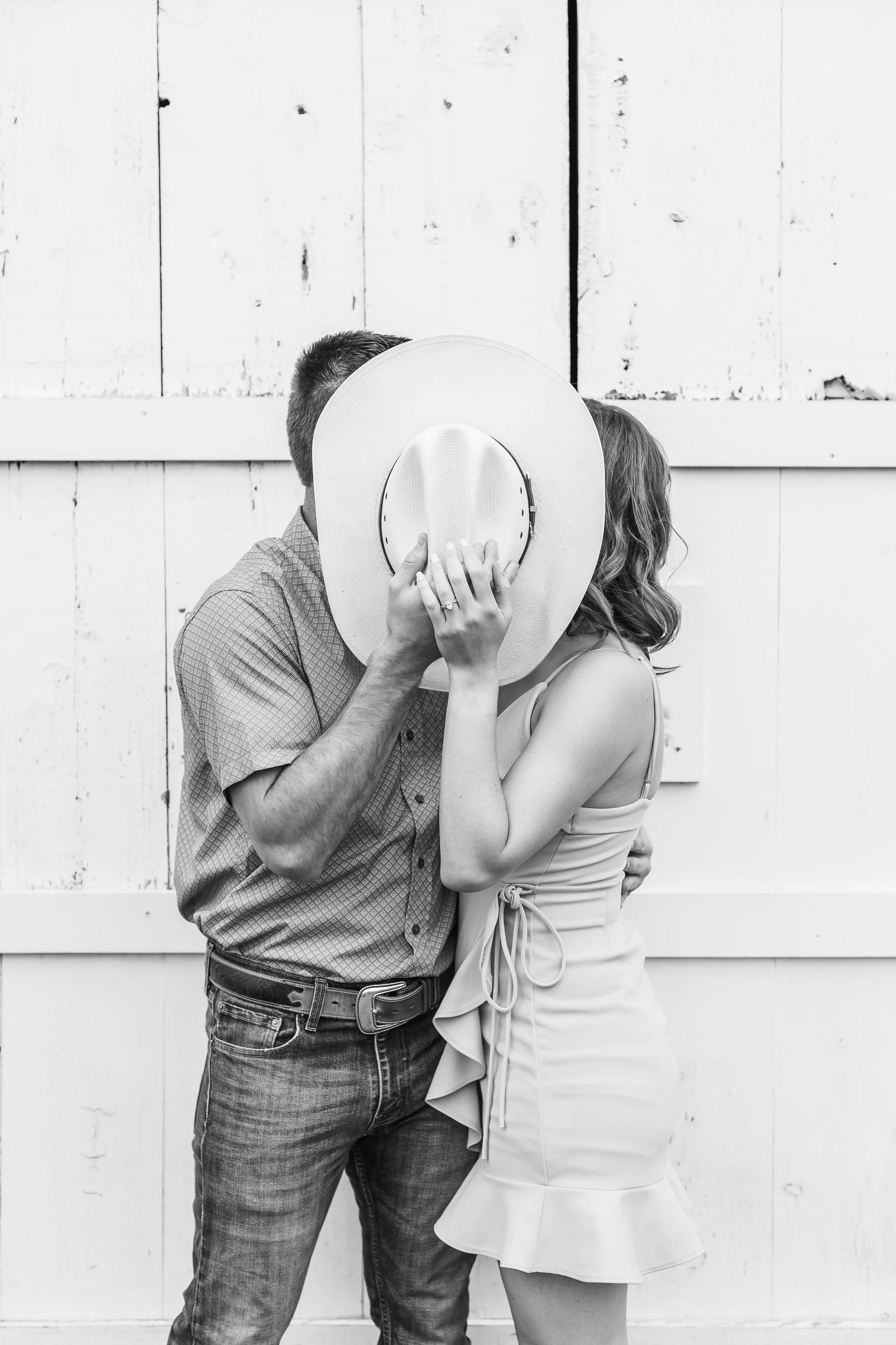 A couple sharing a kiss with a wide-brimmed hat covering their faces, standing in front of a rustic white wooden wall.