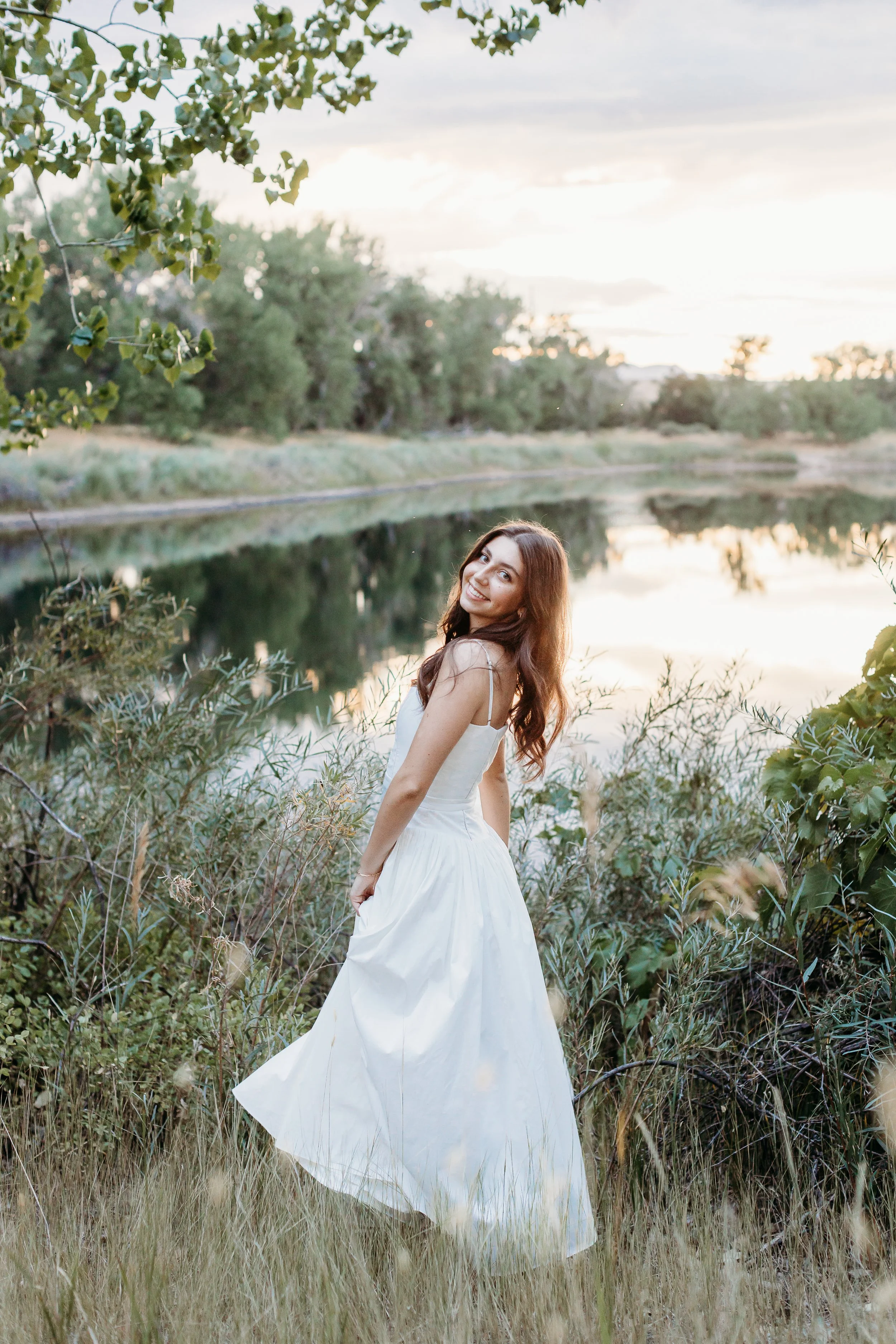 A woman in a white dress standing outdoors by a river at sunset, smiling and looking back at the camera surrounded by greenery.