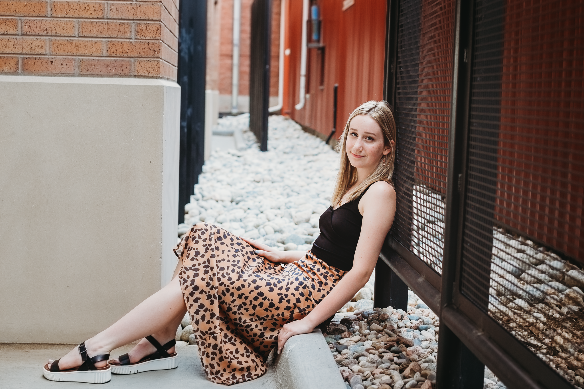 Young woman with blonde hair sitting on a concrete ledge beside a metal fence, smiling at the camera, wearing a black top and leopard-print skirt, with black sandals, outdoors in an alleyway with rocks on the ground.