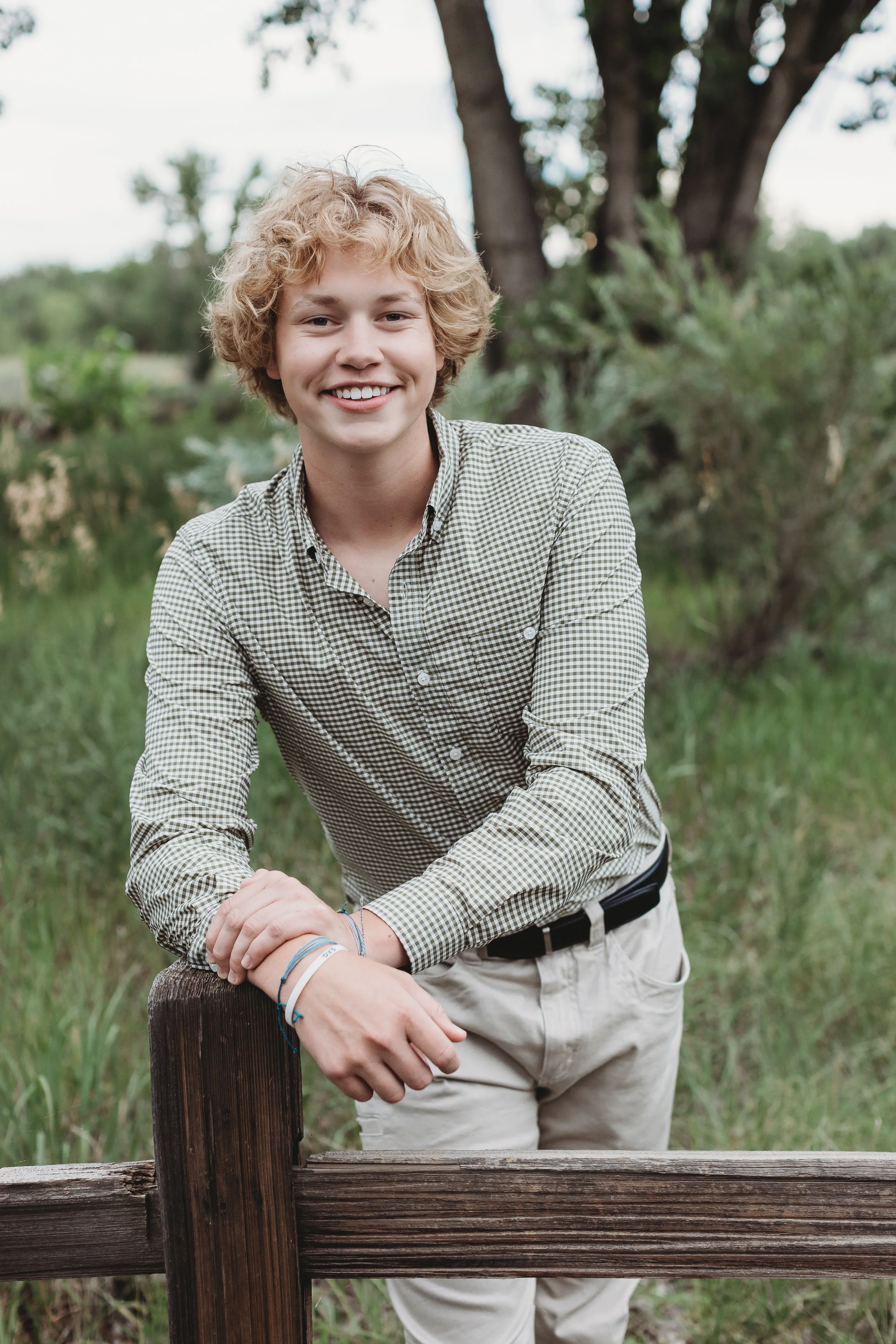 A young man with curly blond hair smiling and leaning on a wooden railing outdoors with trees and grass in the background.