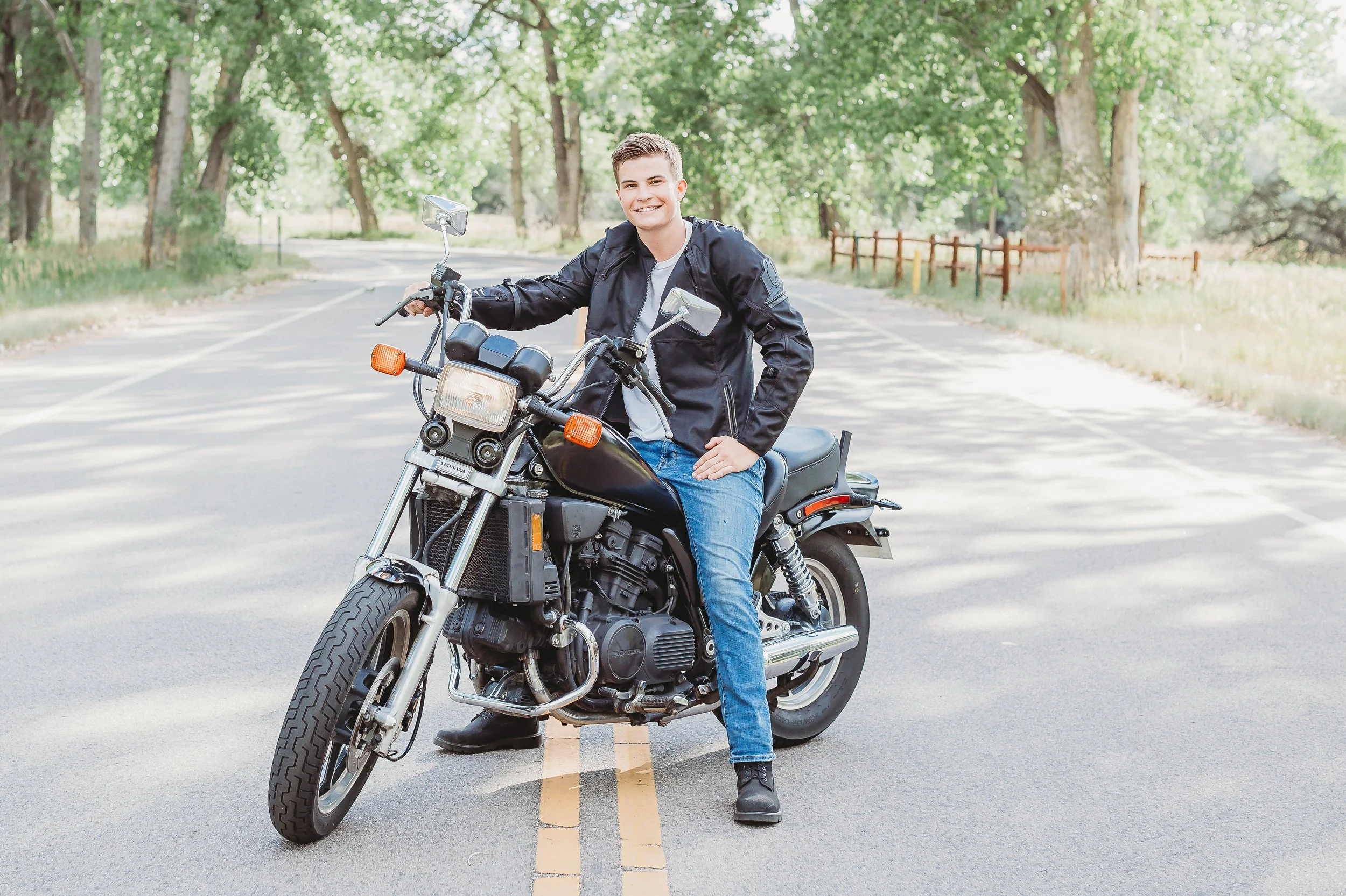 A young man in a black jacket and blue jeans sitting on a black motorcycle in the middle of a tree-lined road, smiling at the camera.