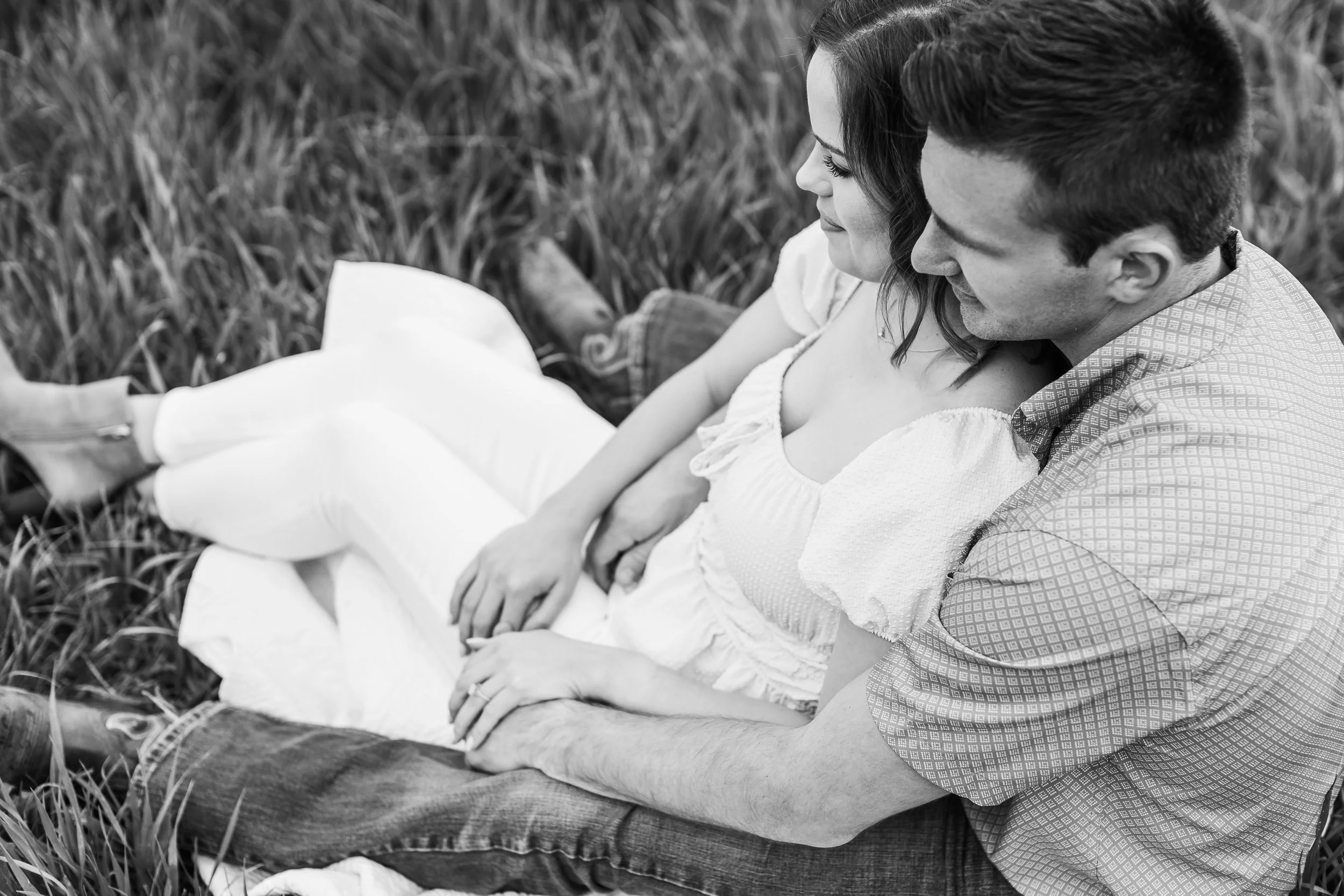 A man and woman sitting closely in a field, holding hands and smiling at each other, in a black-and-white photo.