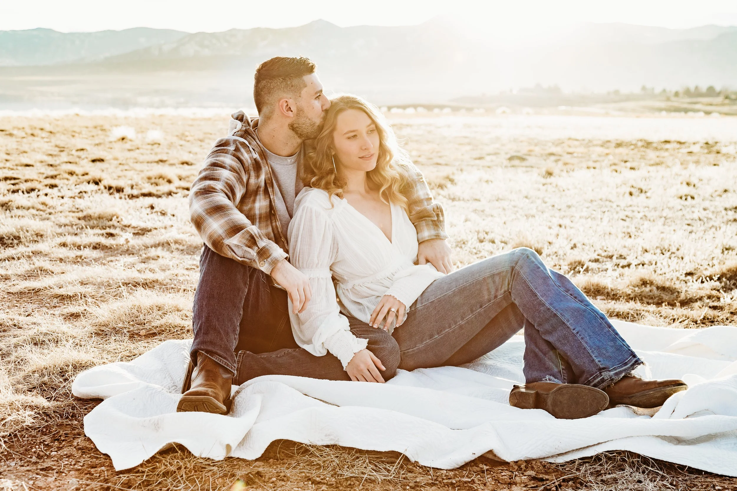 A couple sitting on a blanket in a field near mountains during sunset, with the man kissing the woman on the temple.