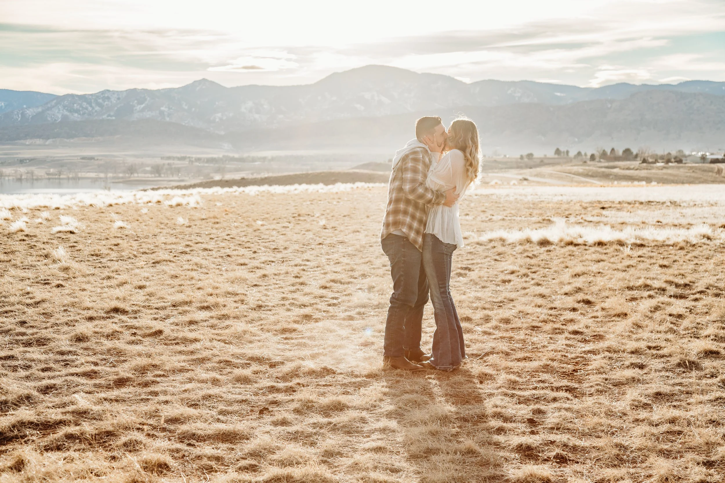 A couple embracing and kissing in a field with mountains in the background during sunset.