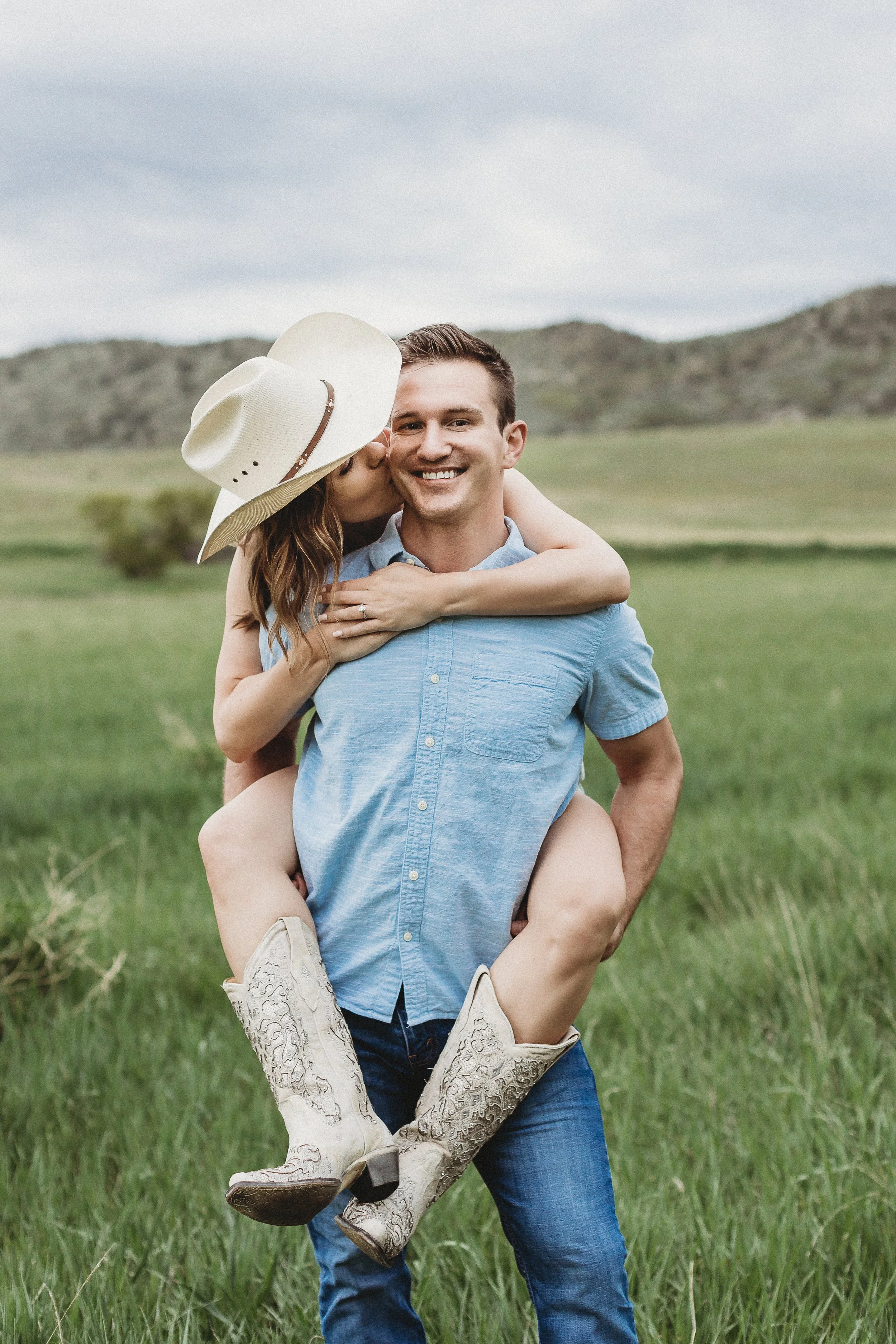A man in a blue shirt giving a piggyback ride to a woman wearing cowboy boots and a large hat, in a green field with hills in the background.
