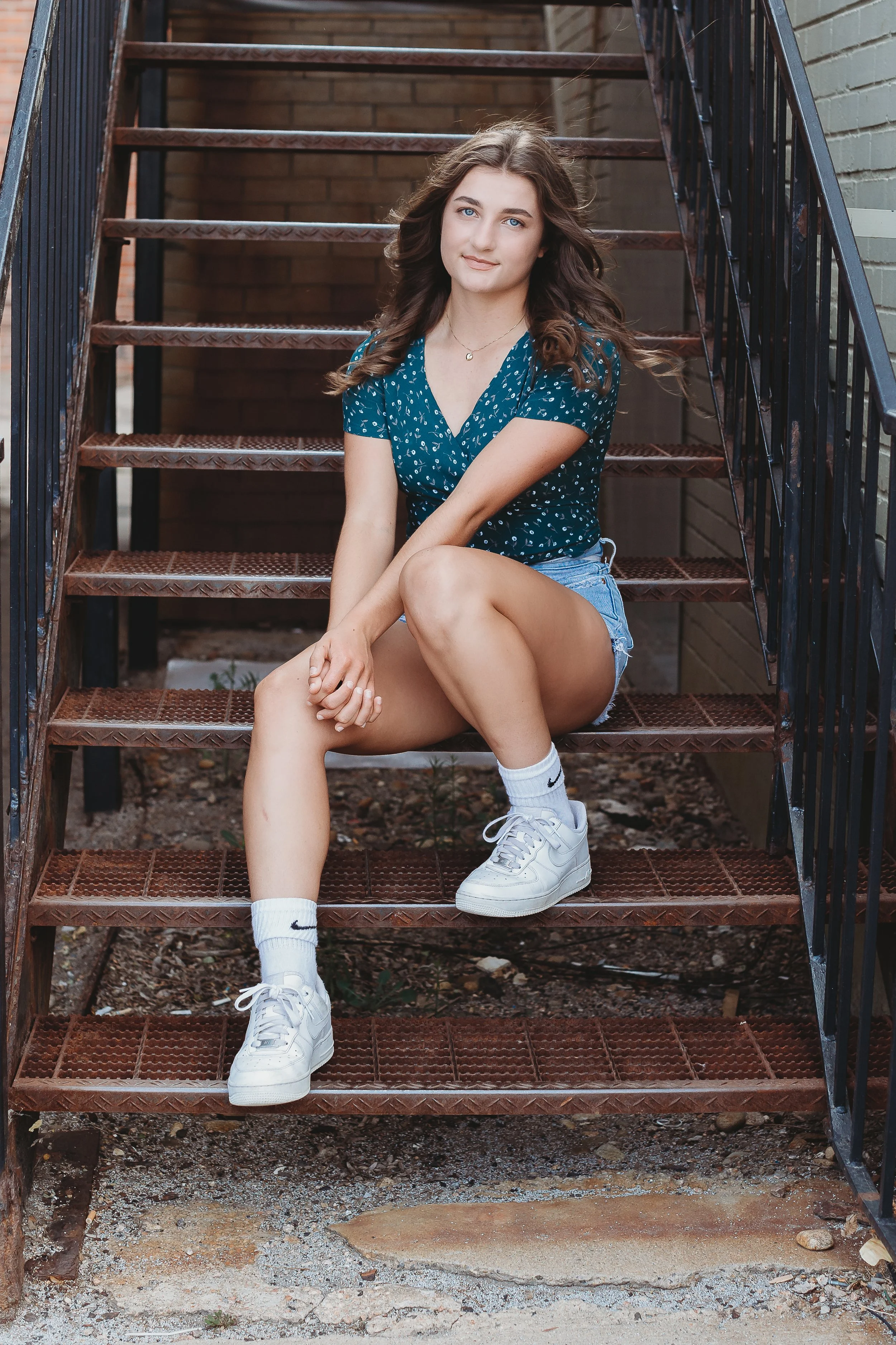 A young woman with long brown hair, sitting on metal outdoor stairs, wearing a blue patterned shirt, denim shorts, white Nike sneakers, and white Nike socks, with a neutral expression.