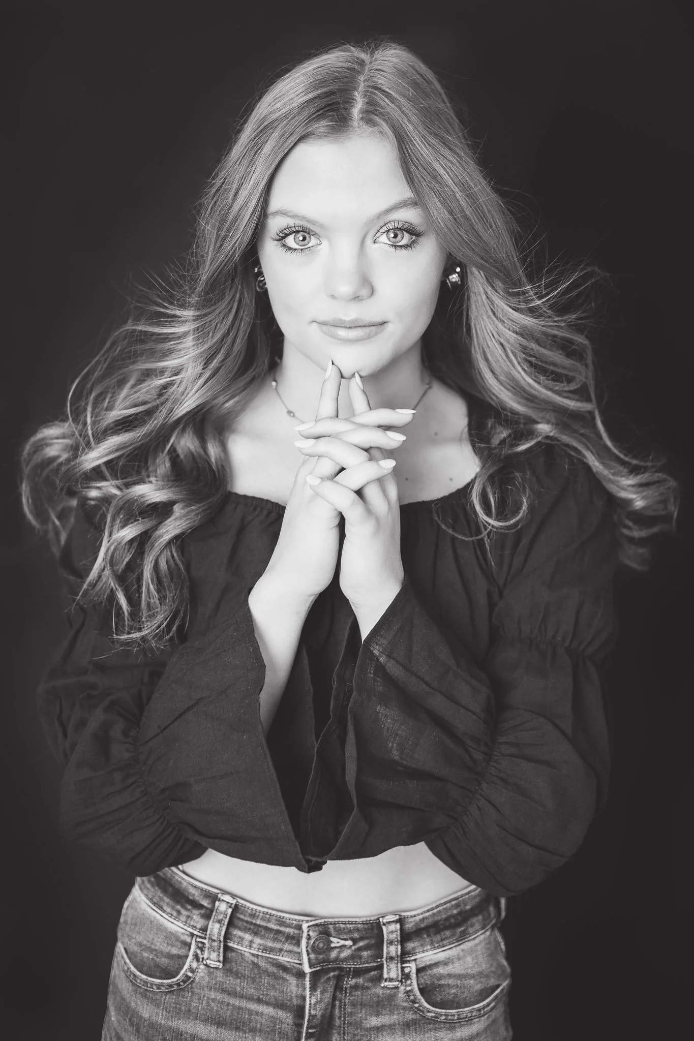 Black and white portrait of a young woman with long, wavy hair, hands clasped near her chin, wearing a dark off-the-shoulder top and denim jeans, looking directly at the camera.