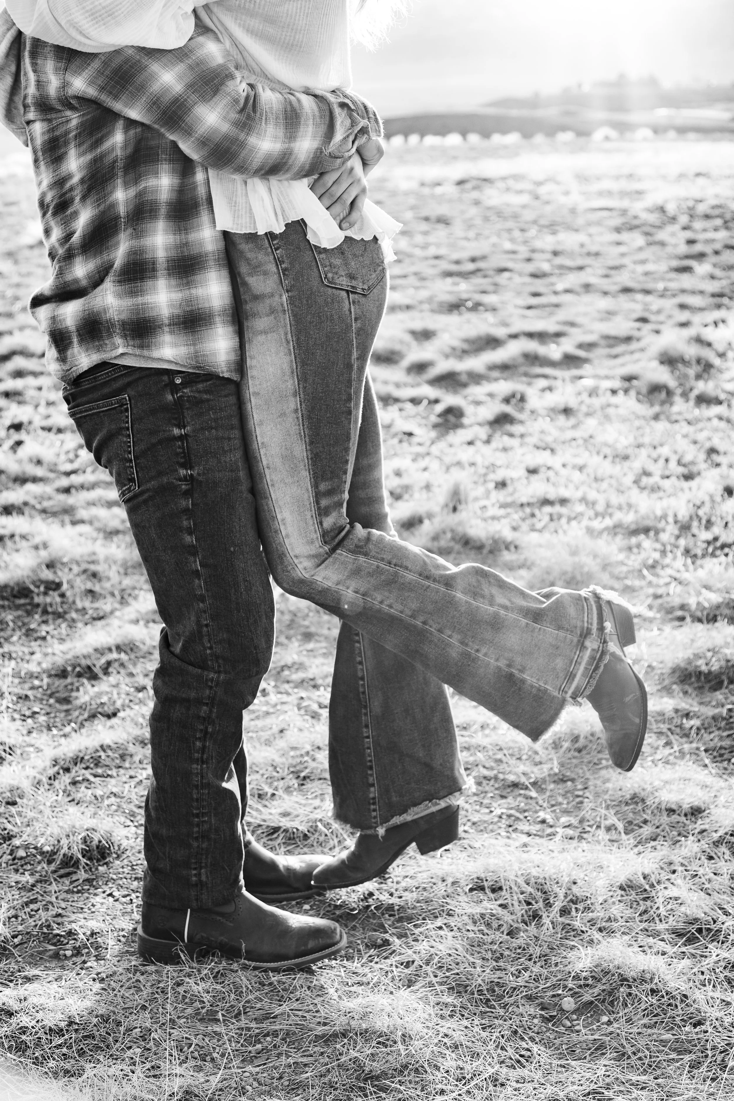 Black and white photo of a couple standing on grass, with their legs intertwined and arms around each other, outdoors.