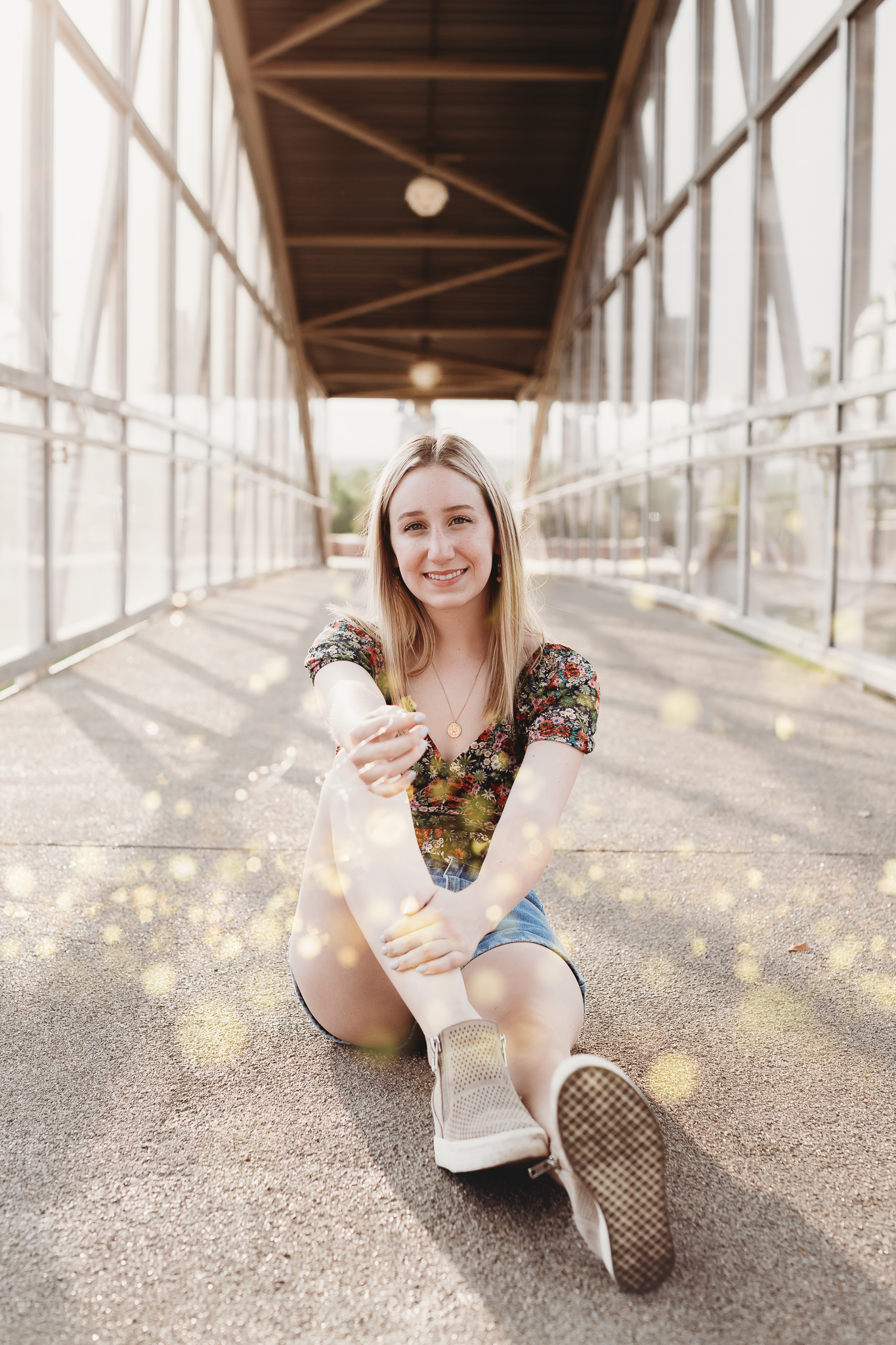 A young woman with blonde hair and a floral shirt sitting on the ground in an enclosed walkway, smiling at the camera, with sunlight and bokeh effects around her.