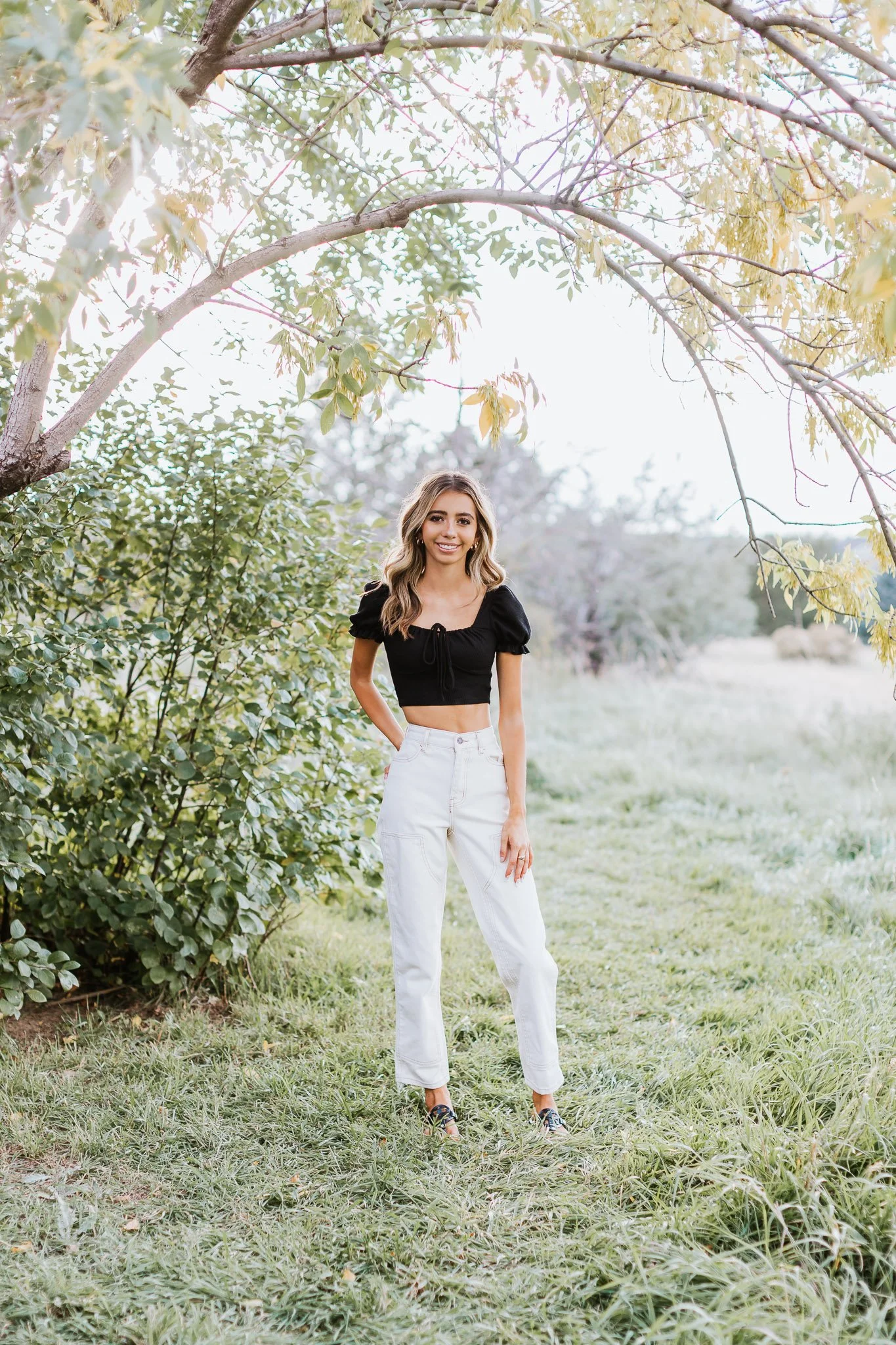 A young woman standing outdoors on a grassy path, surrounded by trees and greenery, smiling at the camera.