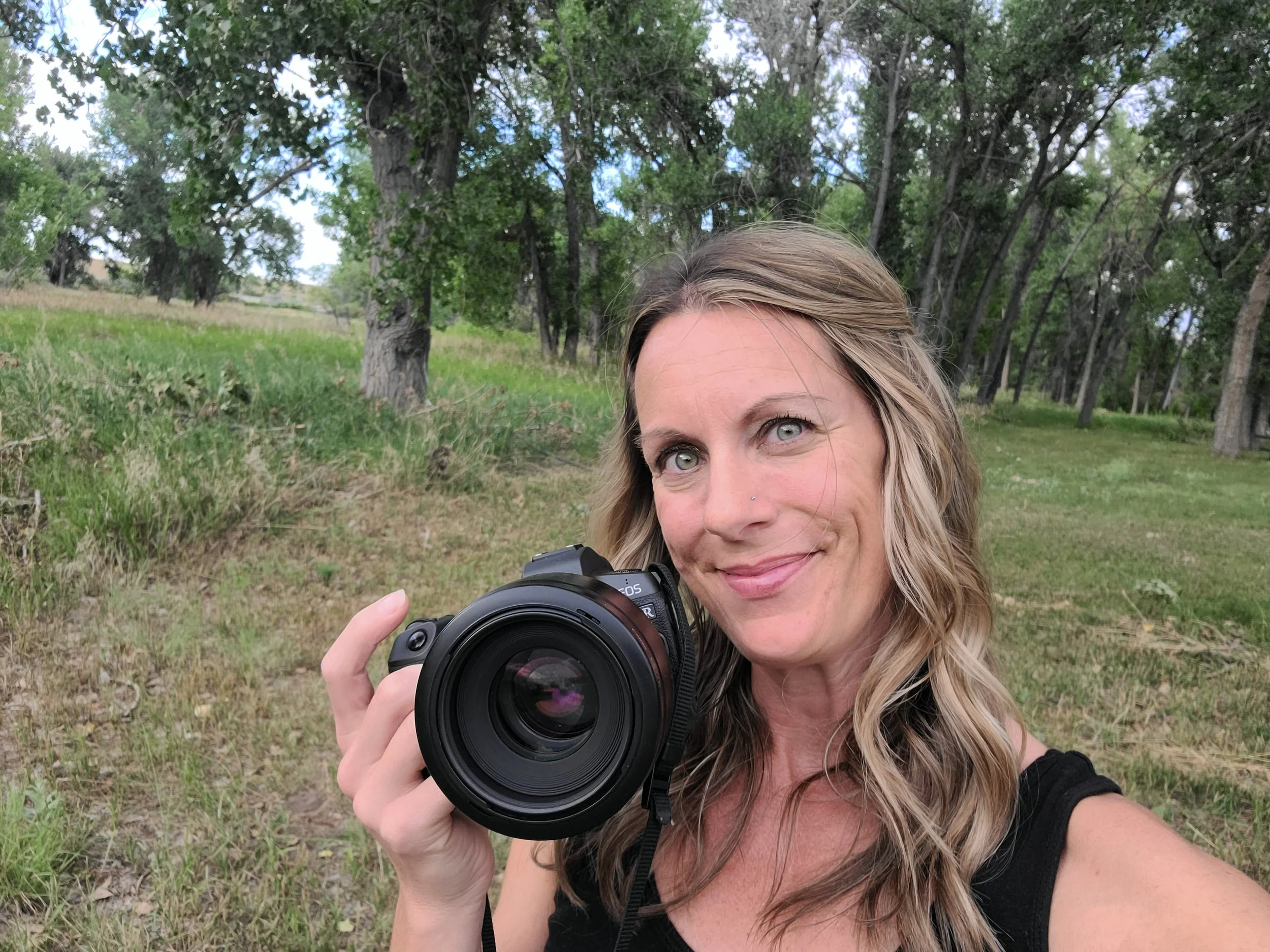 A woman with wavy blonde hair holding a camera outdoors in a grassy, wooded area with trees in the background