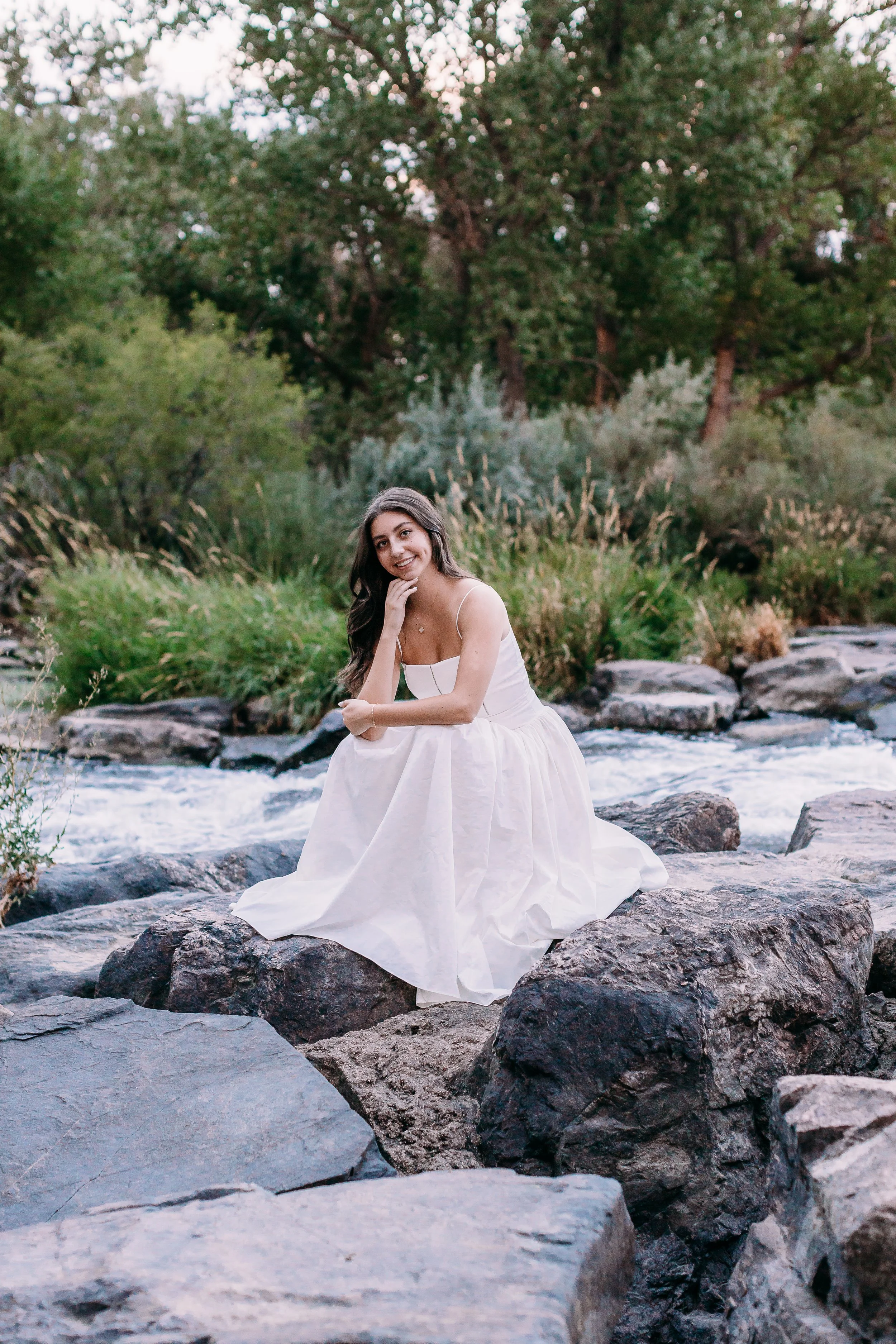 A young woman in a white dress sitting on rocks by a stream in a forested area, smiling at the camera.
