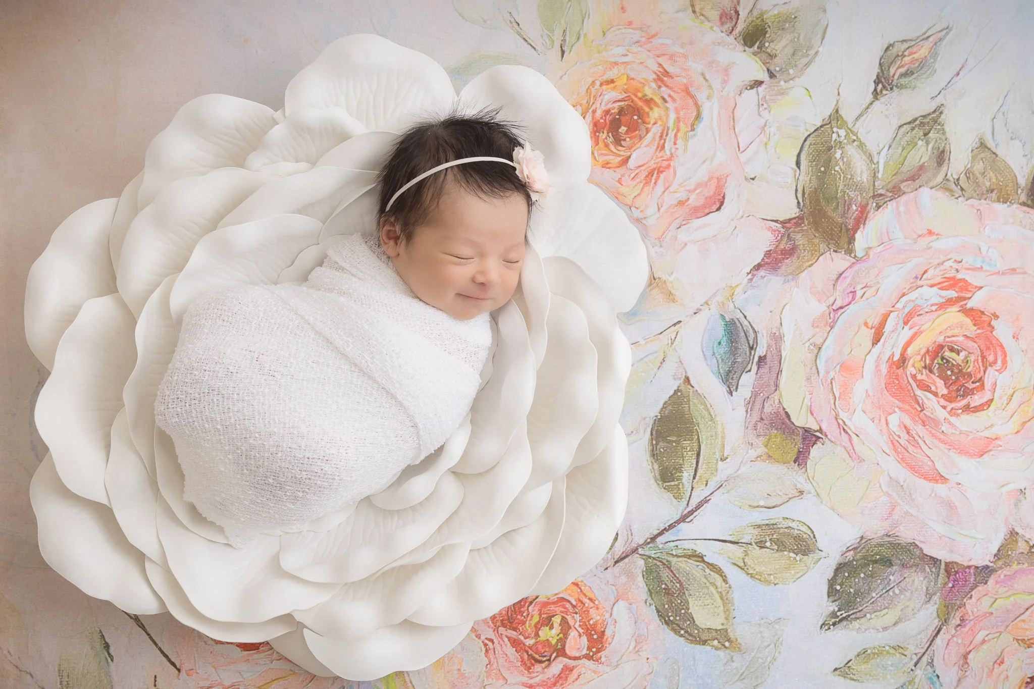 A smiling baby girl wrapped in a white blanket, sleeping on a petal-shaped cushion, with a floral painting background.