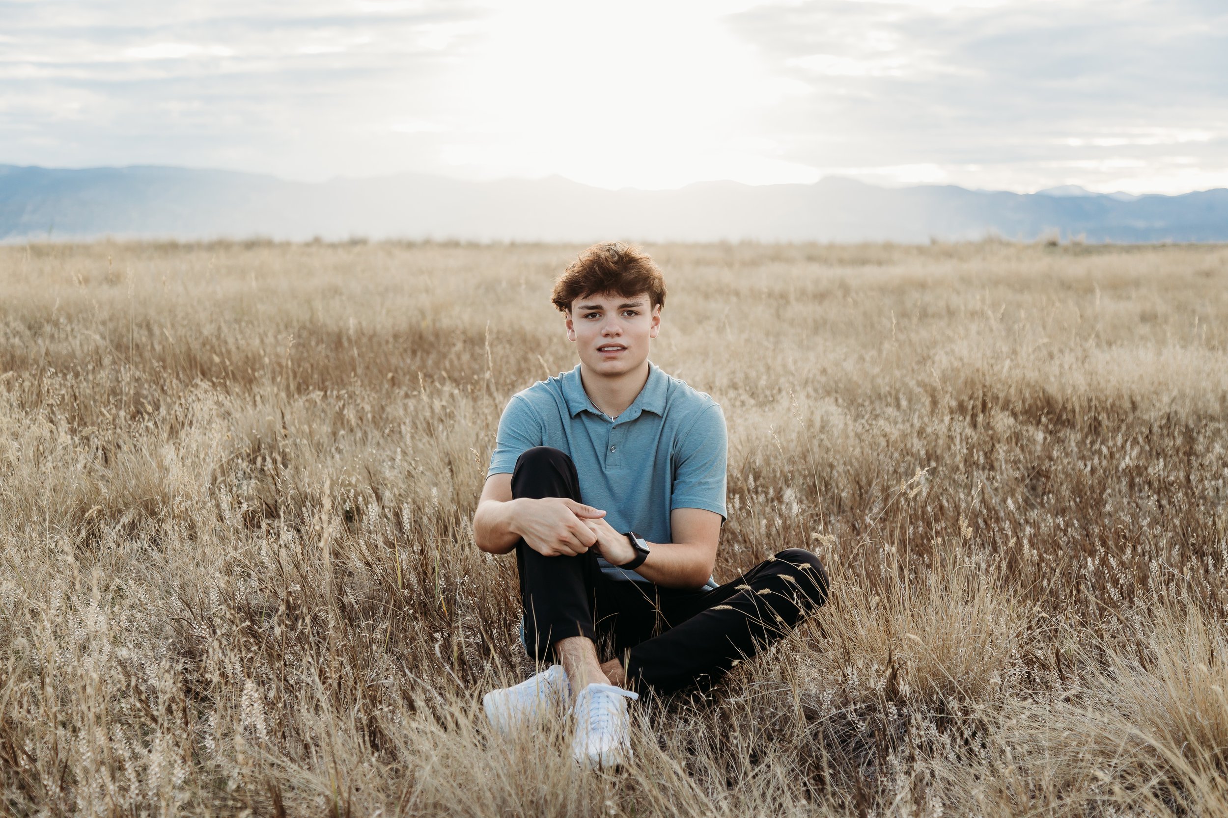 A young man sitting in a field of tall, dry grass with mountains and a cloudy sky in the background.