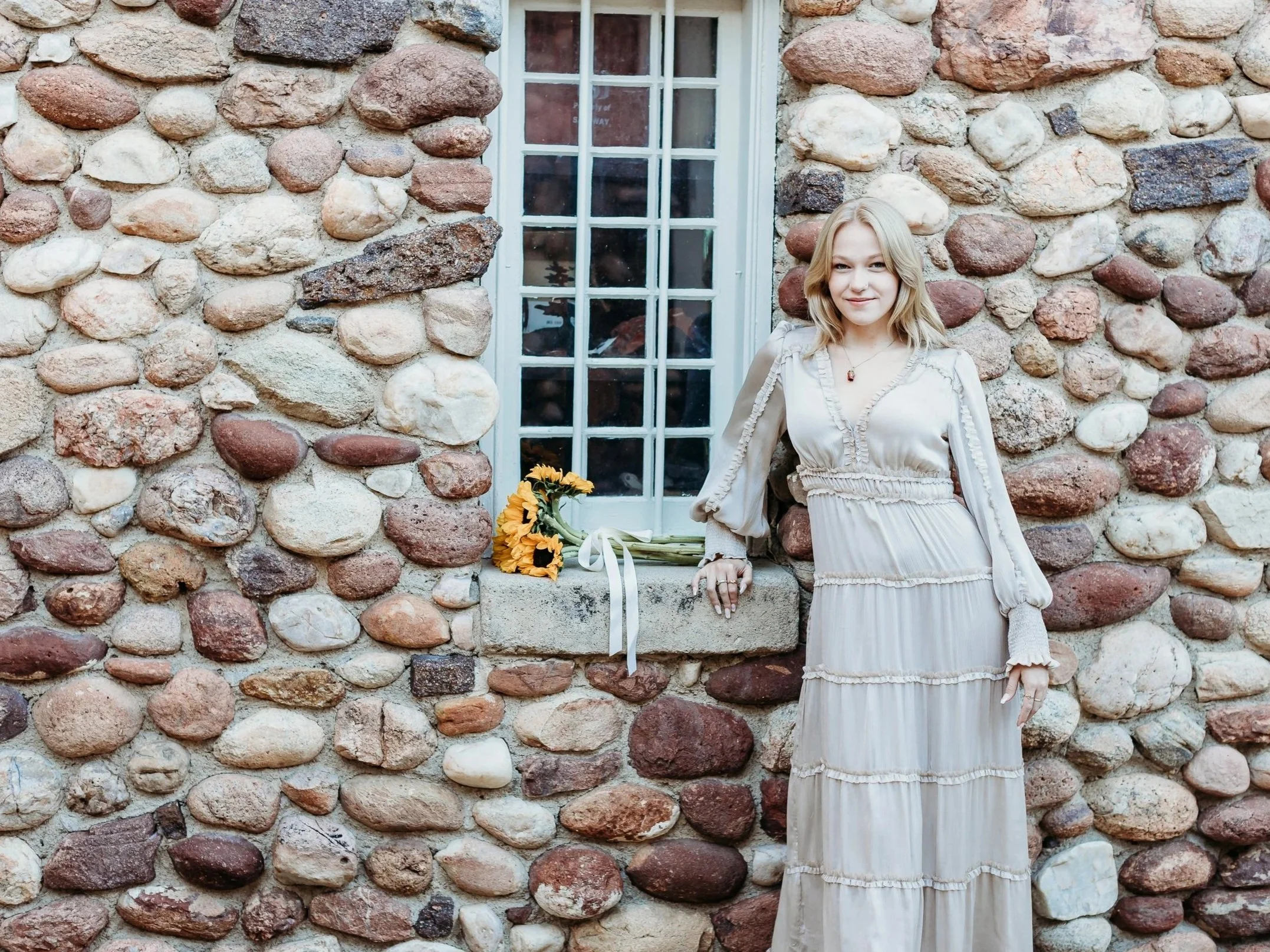 A young woman with shoulder-length blonde hair wearing a long, beige, tiered dress with ruffles stands outside a stone building, next to a window with a bouquet of sunflowers on the window ledge.