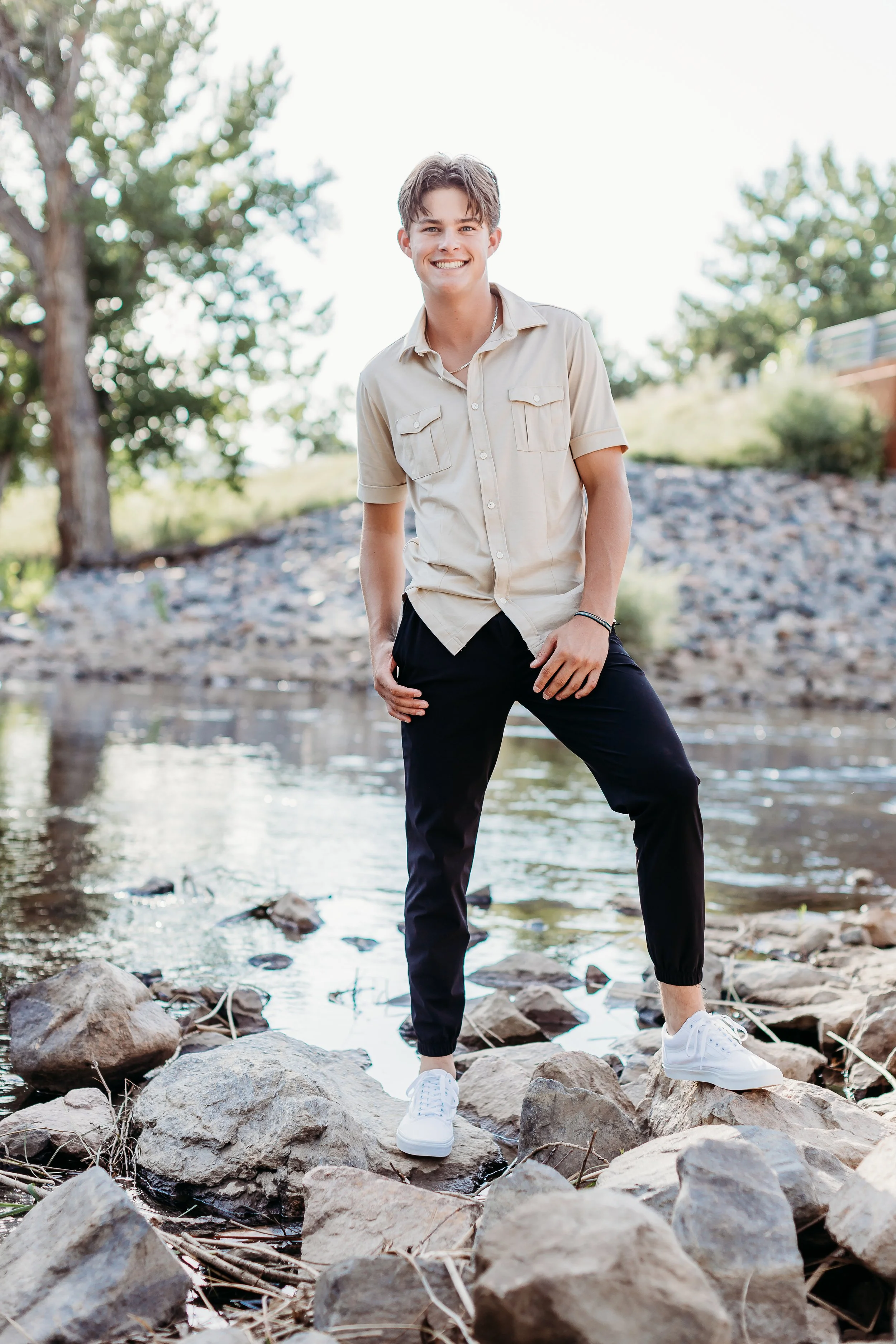 Young man standing on rocks near a creek, outdoors, smiling, wearing a beige shirt and black pants.