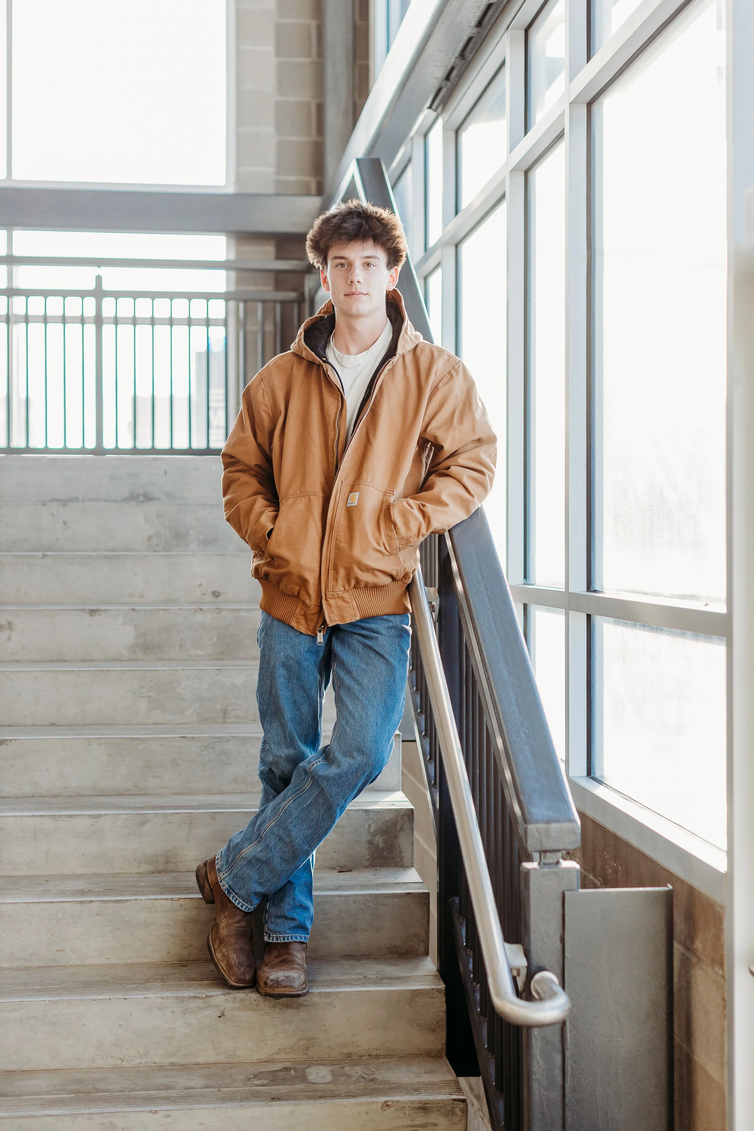 A young man with brown hair standing on a staircase, wearing a brown jacket, blue jeans, and brown boots, leaning against the handrail near large window panels.