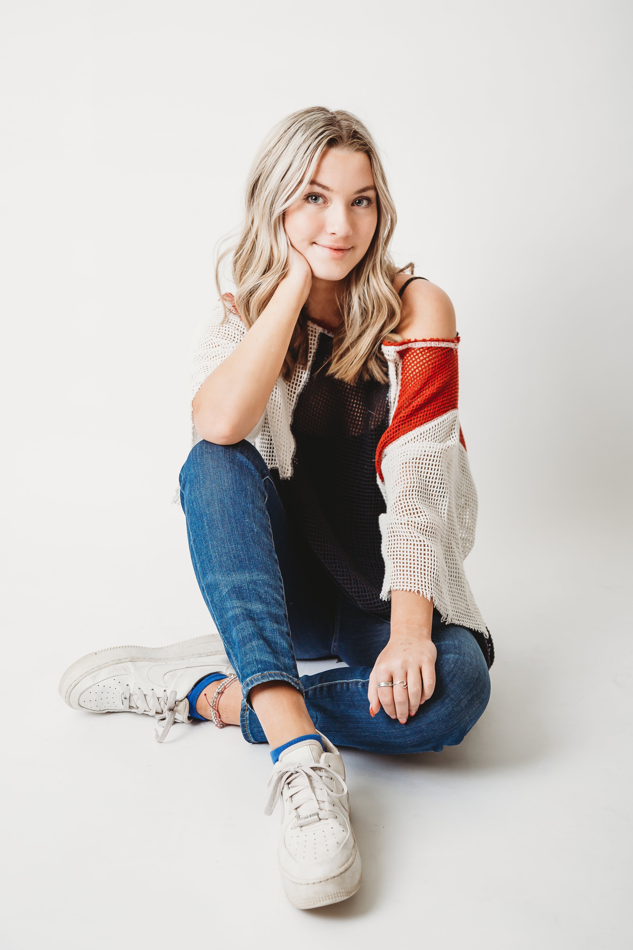 A young woman with blonde hair, wearing a black top, a white and red mesh cardigan, blue jeans, and white sneakers, sitting on the floor with her legs crossed, resting her chin on her hand, and smiling at the camera.