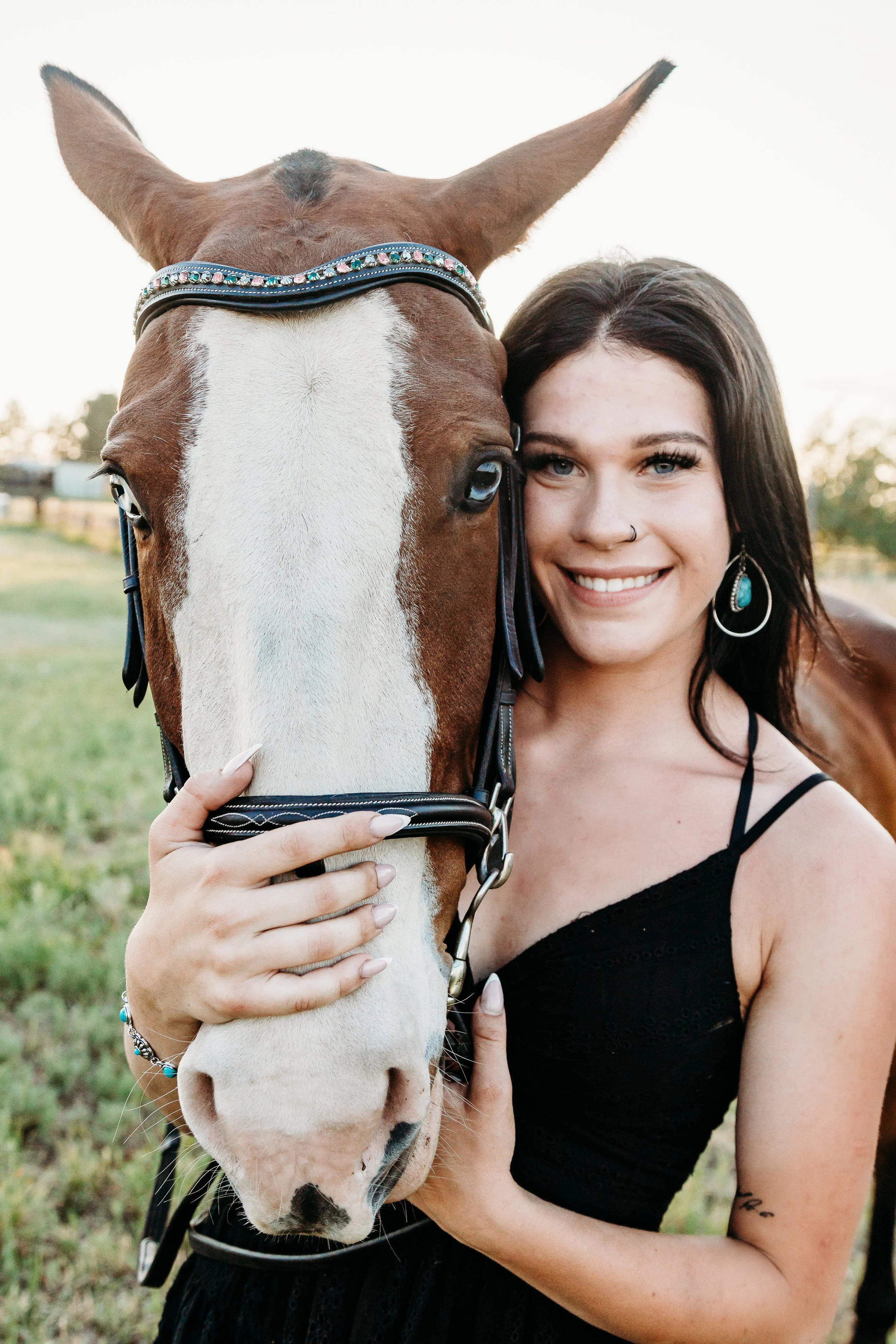 A young woman with dark hair, earrings, and a nose ring, smiling and hugging a brown and white horse with a decorative bridle, outdoors during sunset.