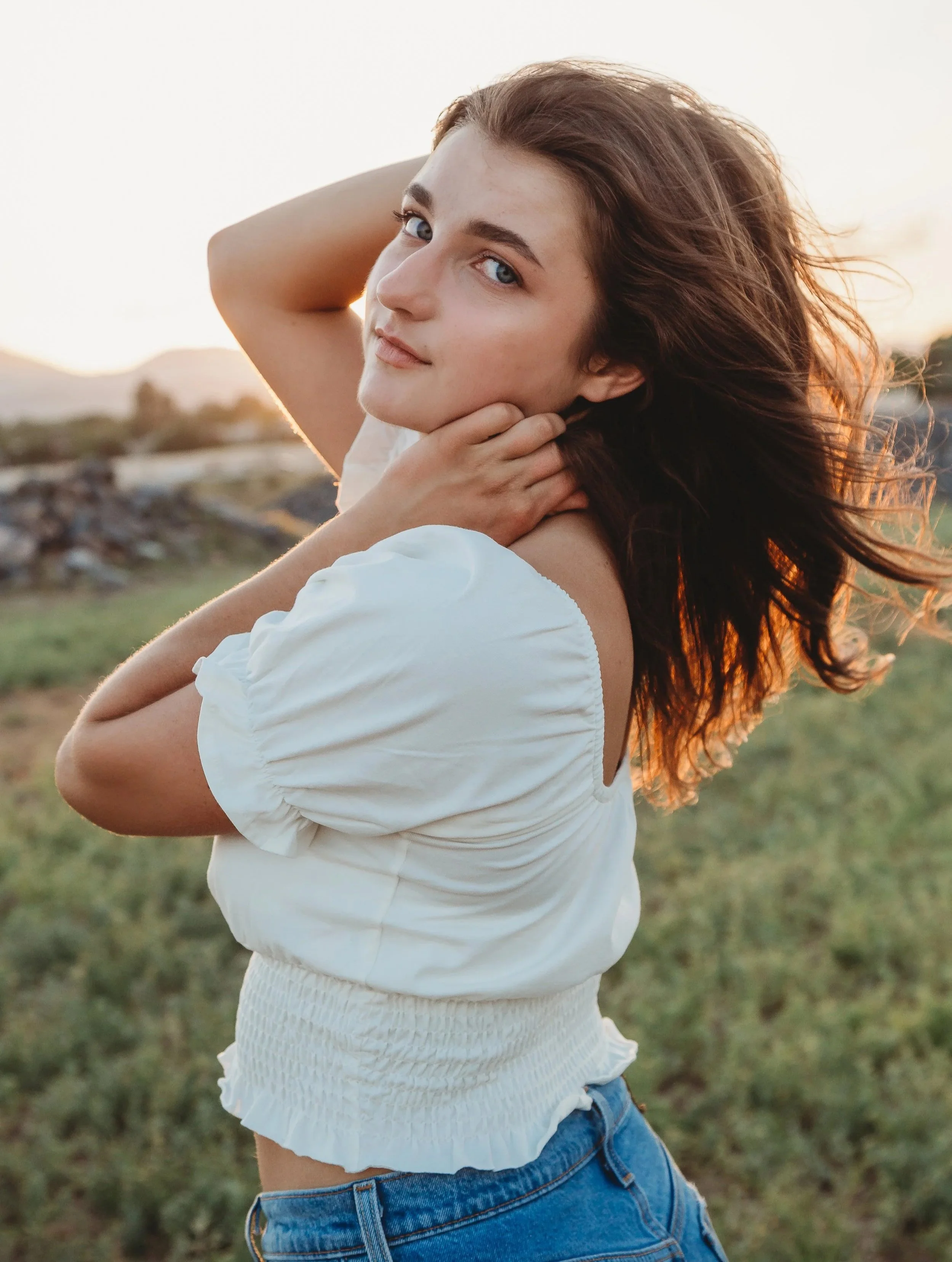 A young woman with brown hair and blue eyes posing outdoors during sunset, wearing a white puff-sleeve top and blue jeans.