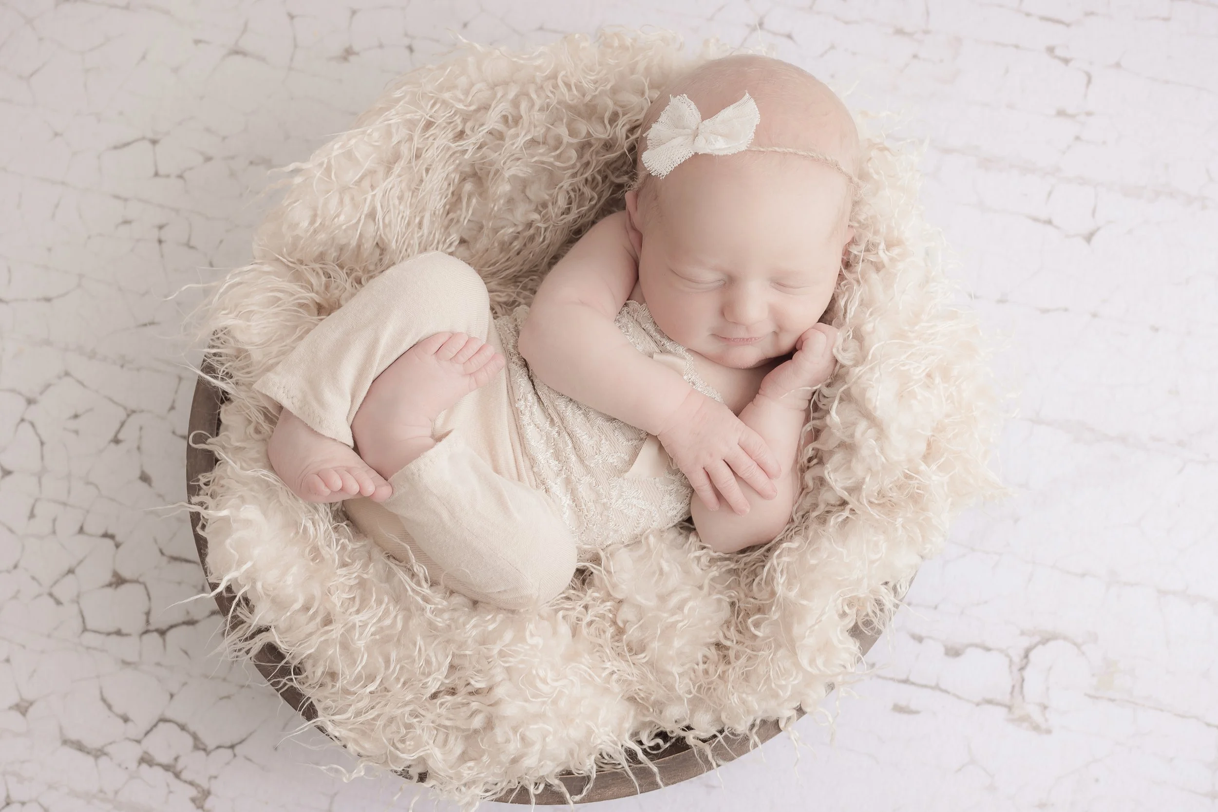 A sleeping baby with a cream-colored headband and a bow, lying on a fluffy beige blanket inside a round basket on a white, cracked surface.
