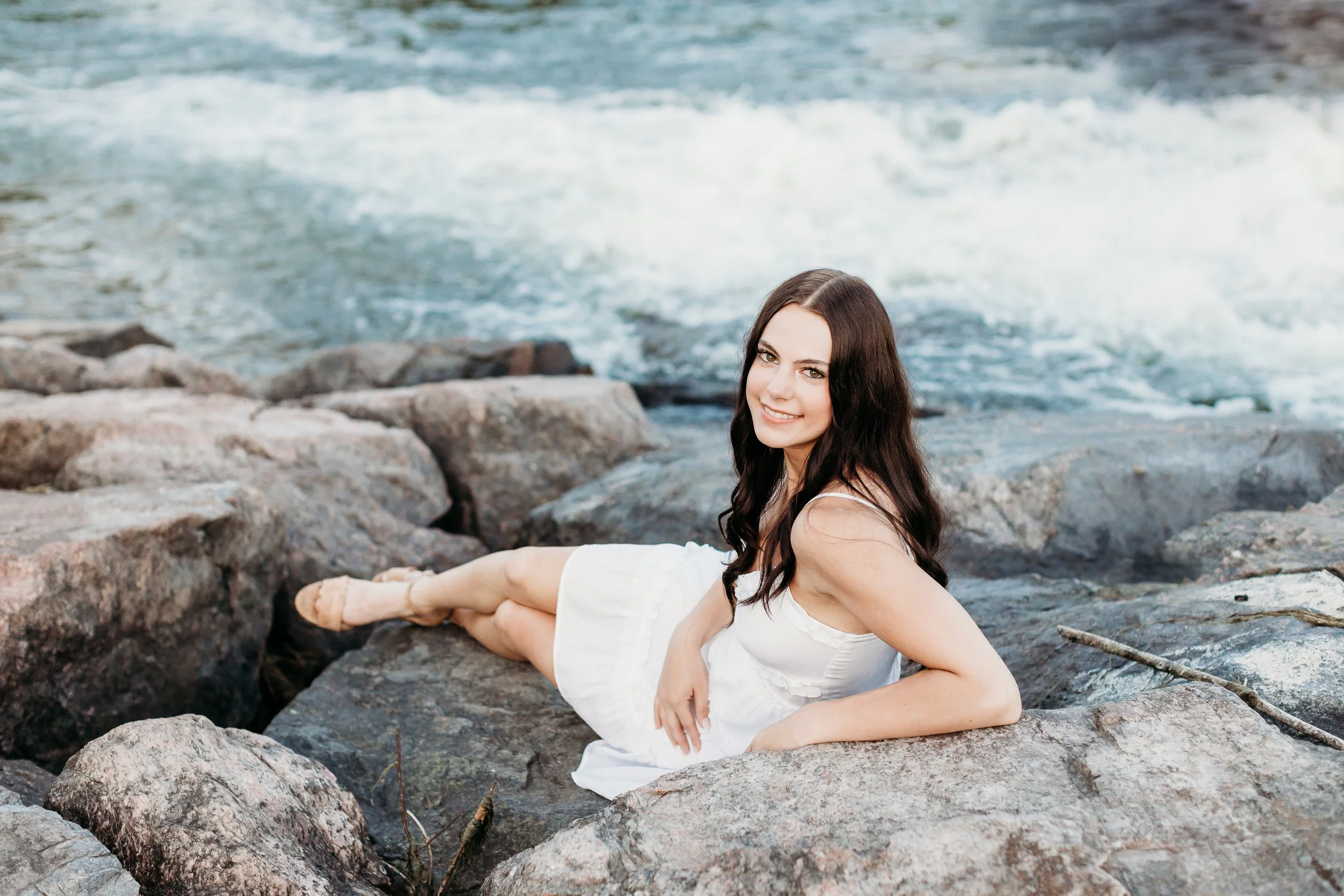 A woman in a white dress lying on rocks by a river with flowing water, smiling at the camera.
