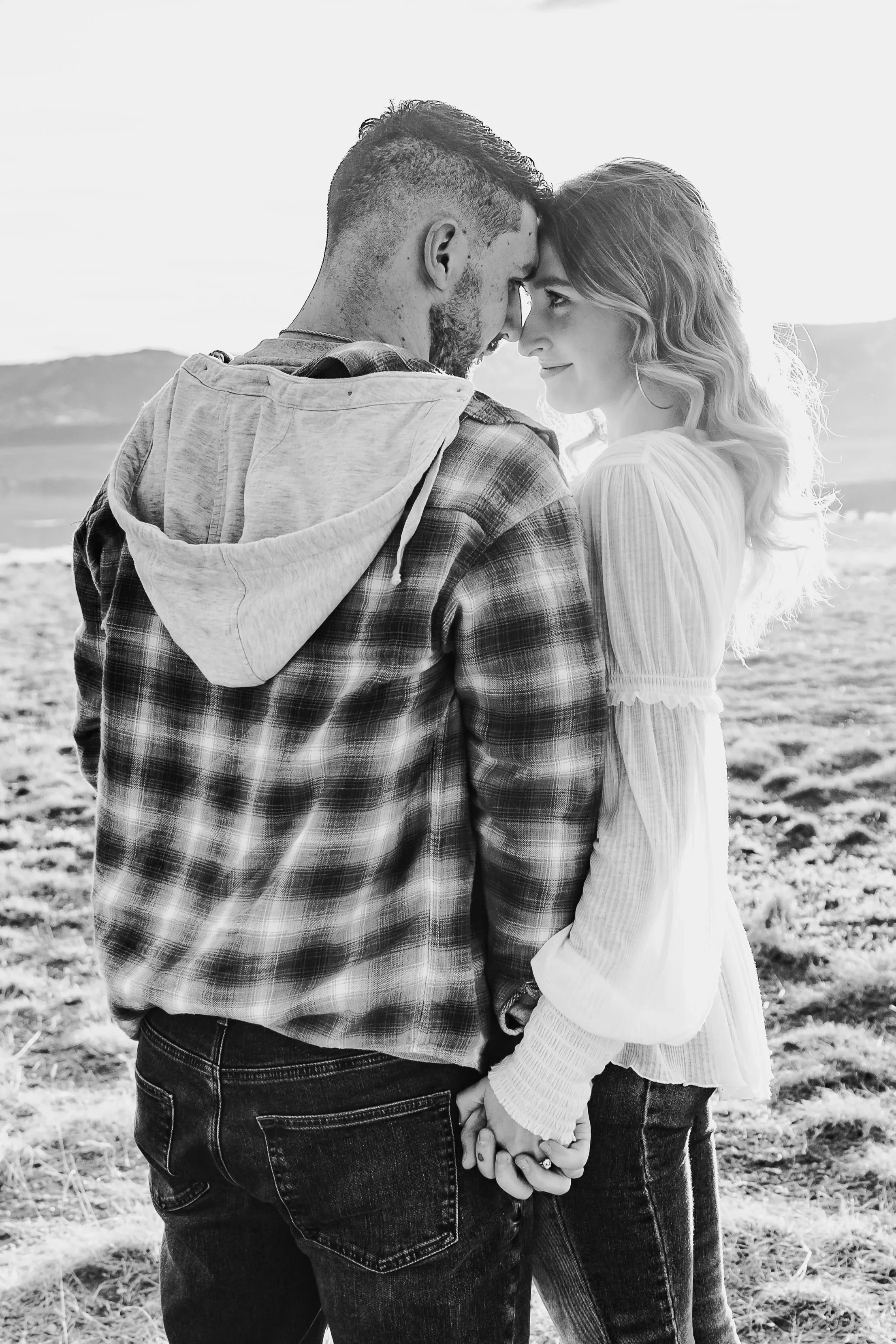 A black-and-white photo of a couple holding hands, with foreheads touching, smiling and looking into each other's eyes with mountains in the background.