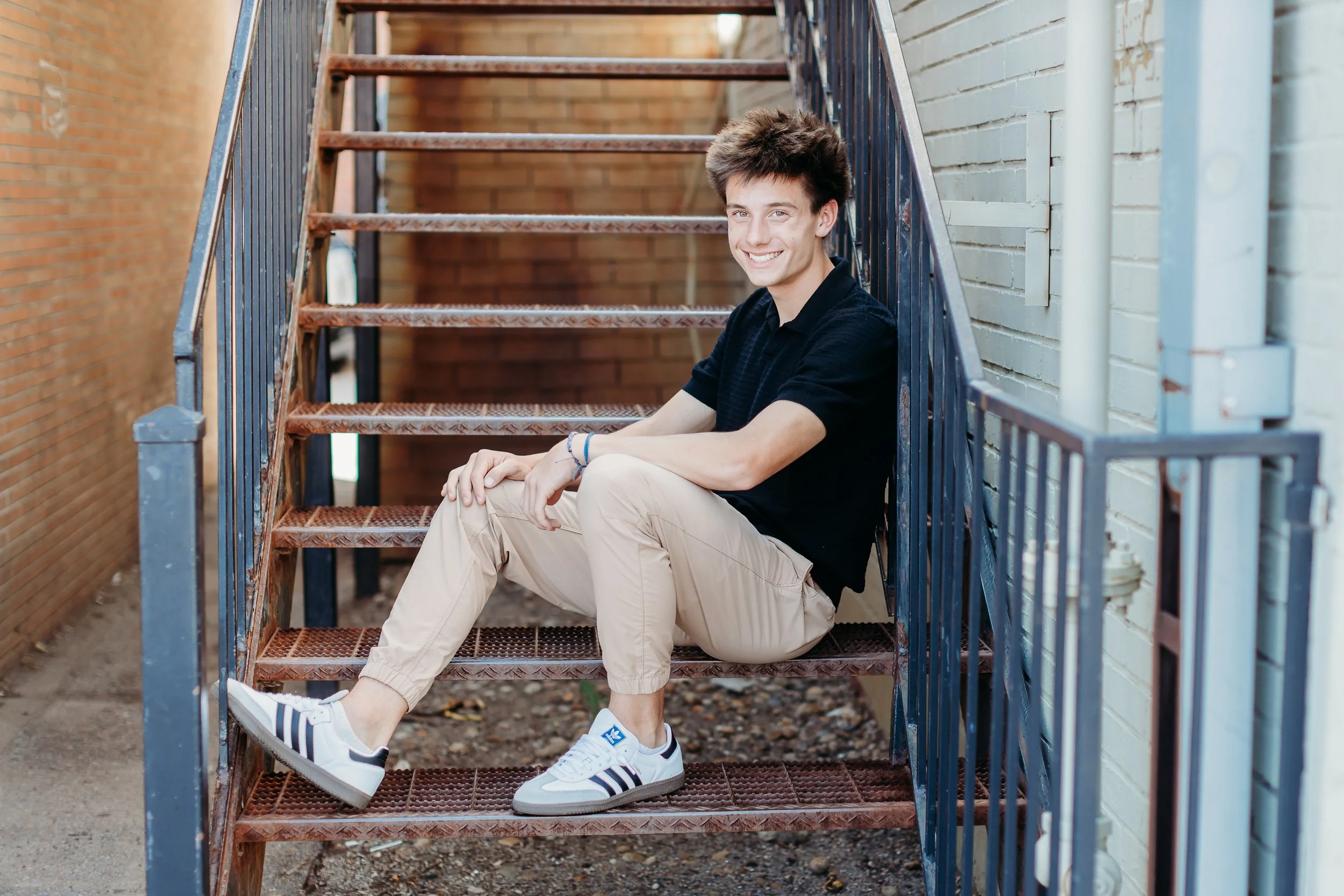 Young man sitting on outdoor metal staircase against brick wall, smiling and looking at camera.