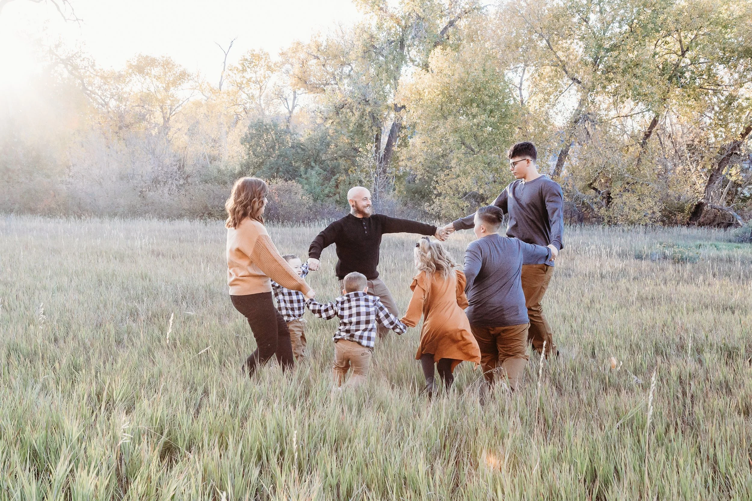 A group of people playing and holding hands in a circle in an open grassy field surrounded by trees during daytime.