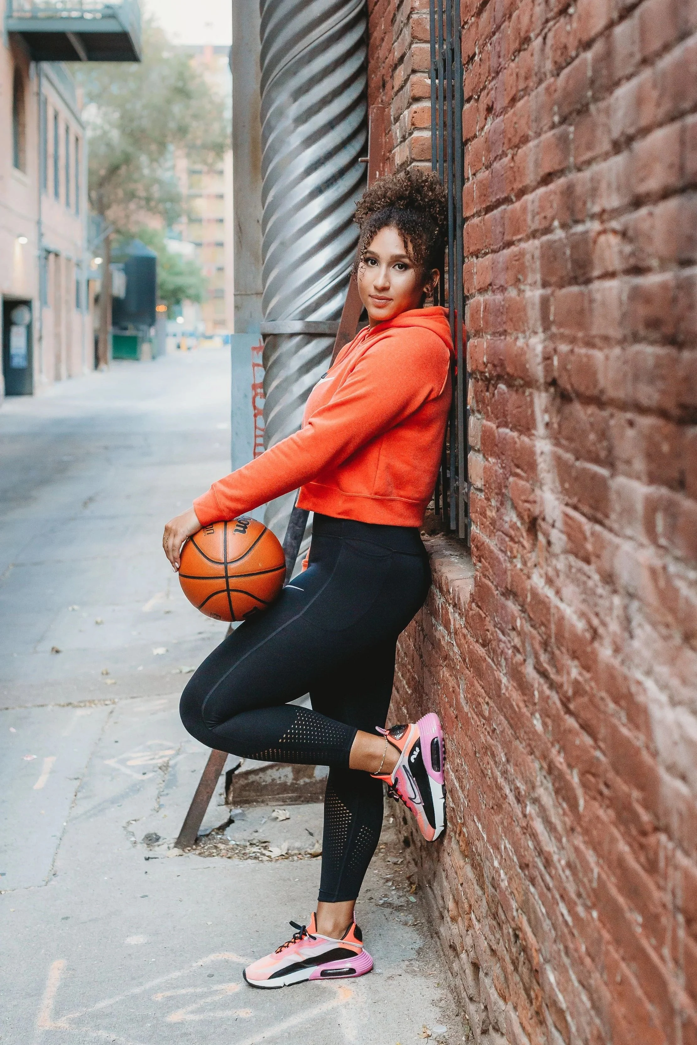 A young woman with curly hair in an orange hoodie and black athletic leggings holding a basketball, leaning against a brick wall in an urban setting.