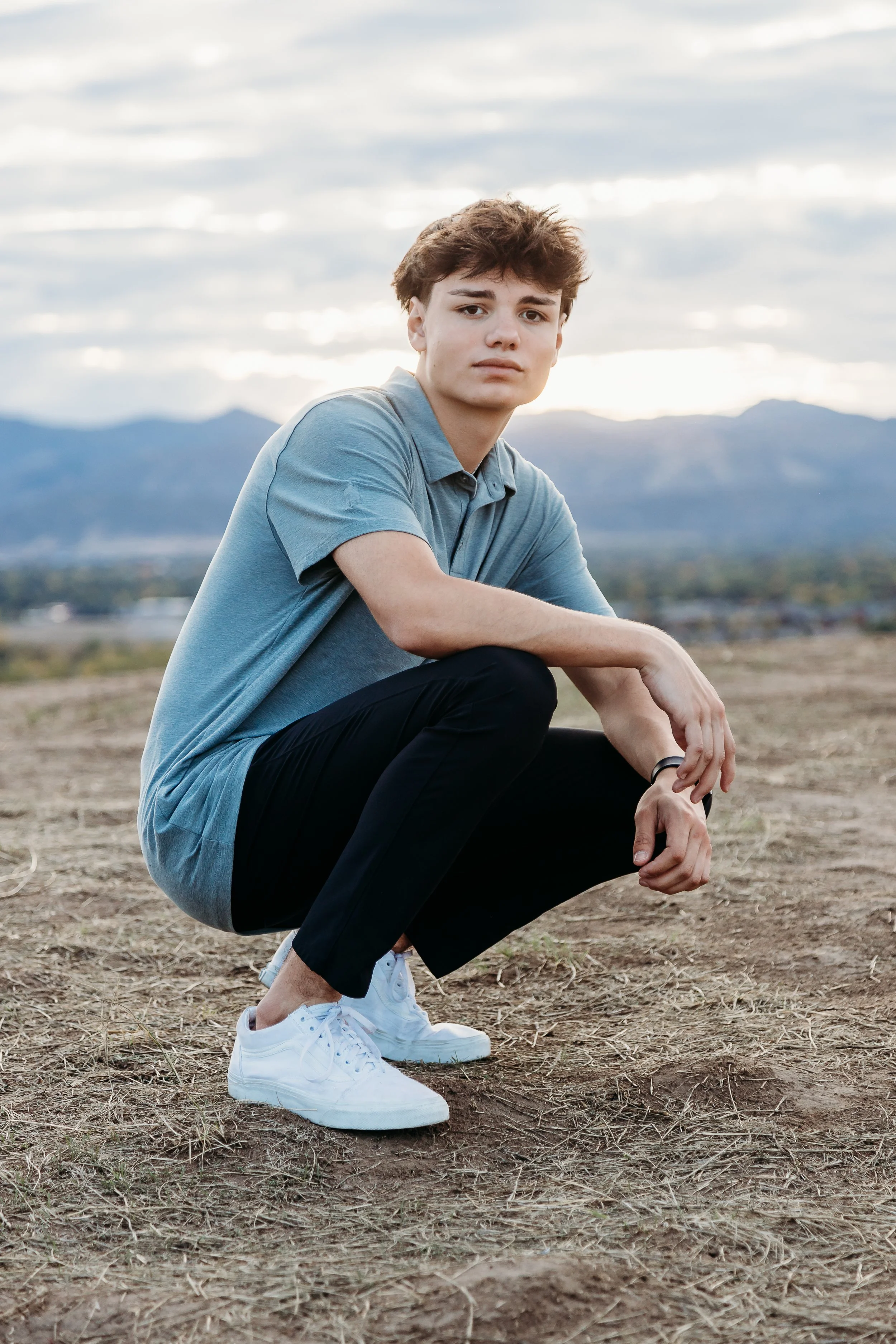 A young man in a gray polo shirt and black pants squatting on dry, grassy ground outdoors with mountains and a cloudy sky in the background.