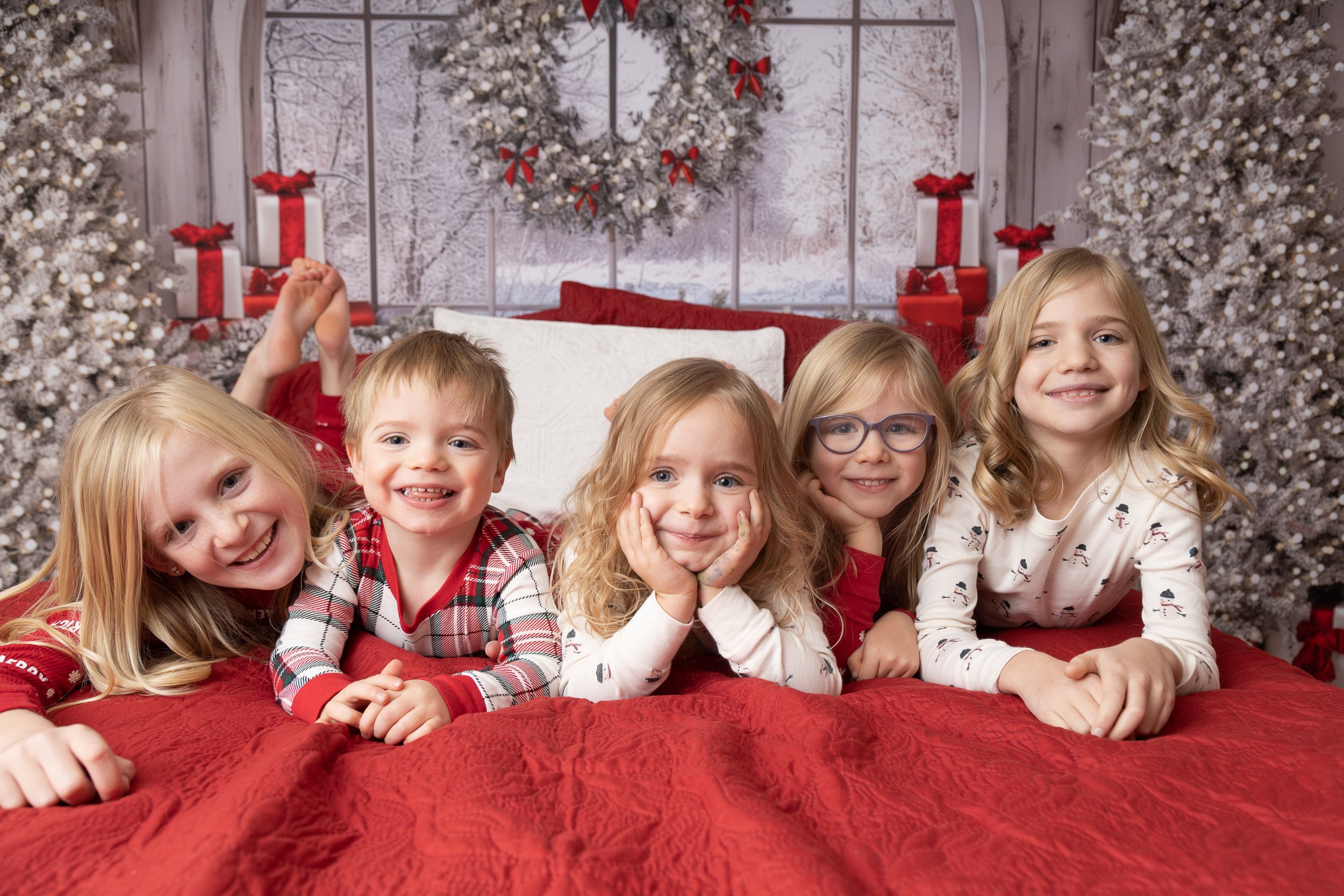 Five children lying on a red bedspread in front of Christmas trees and holiday decorations, smiling and posing for the camera.