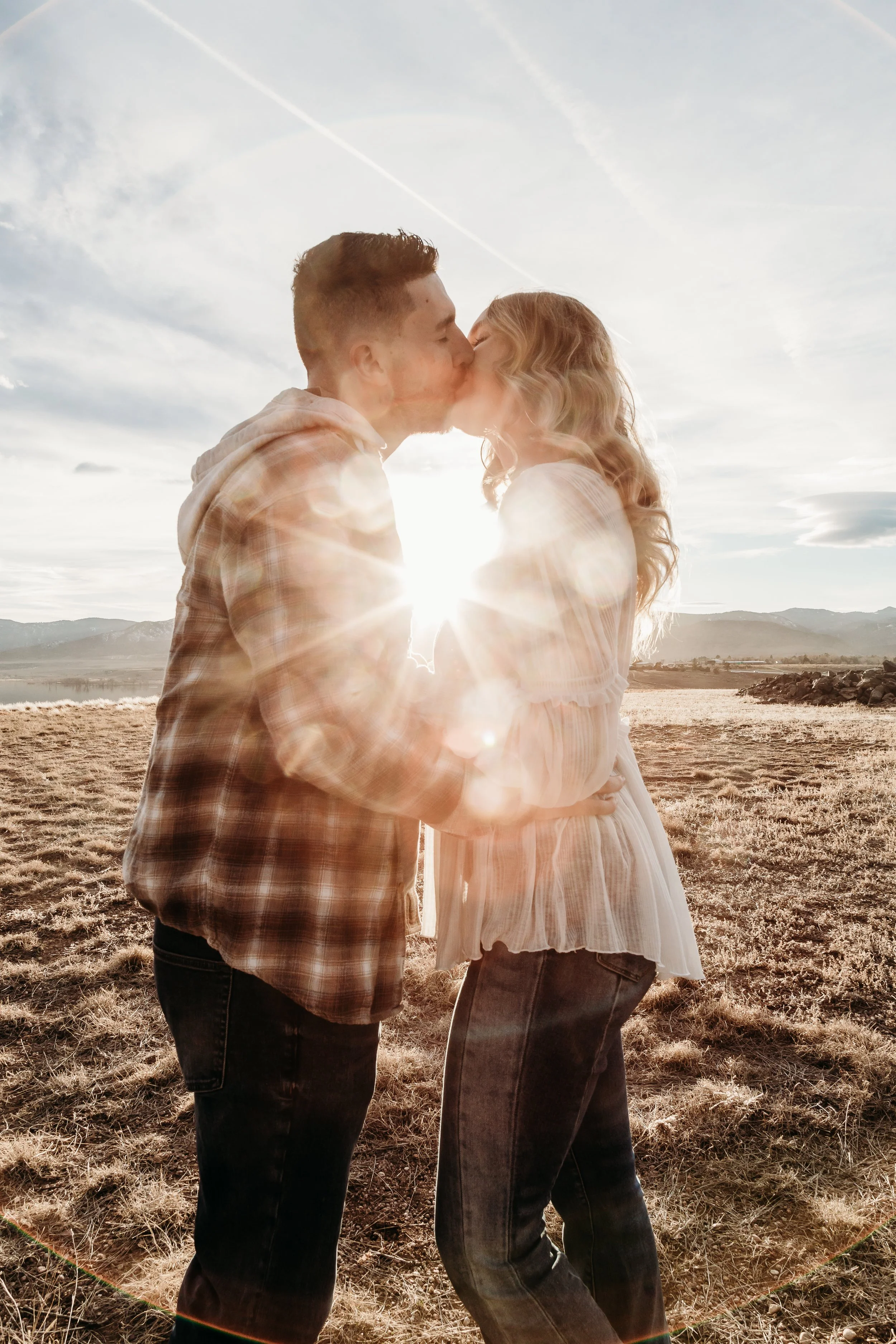 A couple kisses outdoors during sunset, with the sun behind them and mountain scenery in the background.