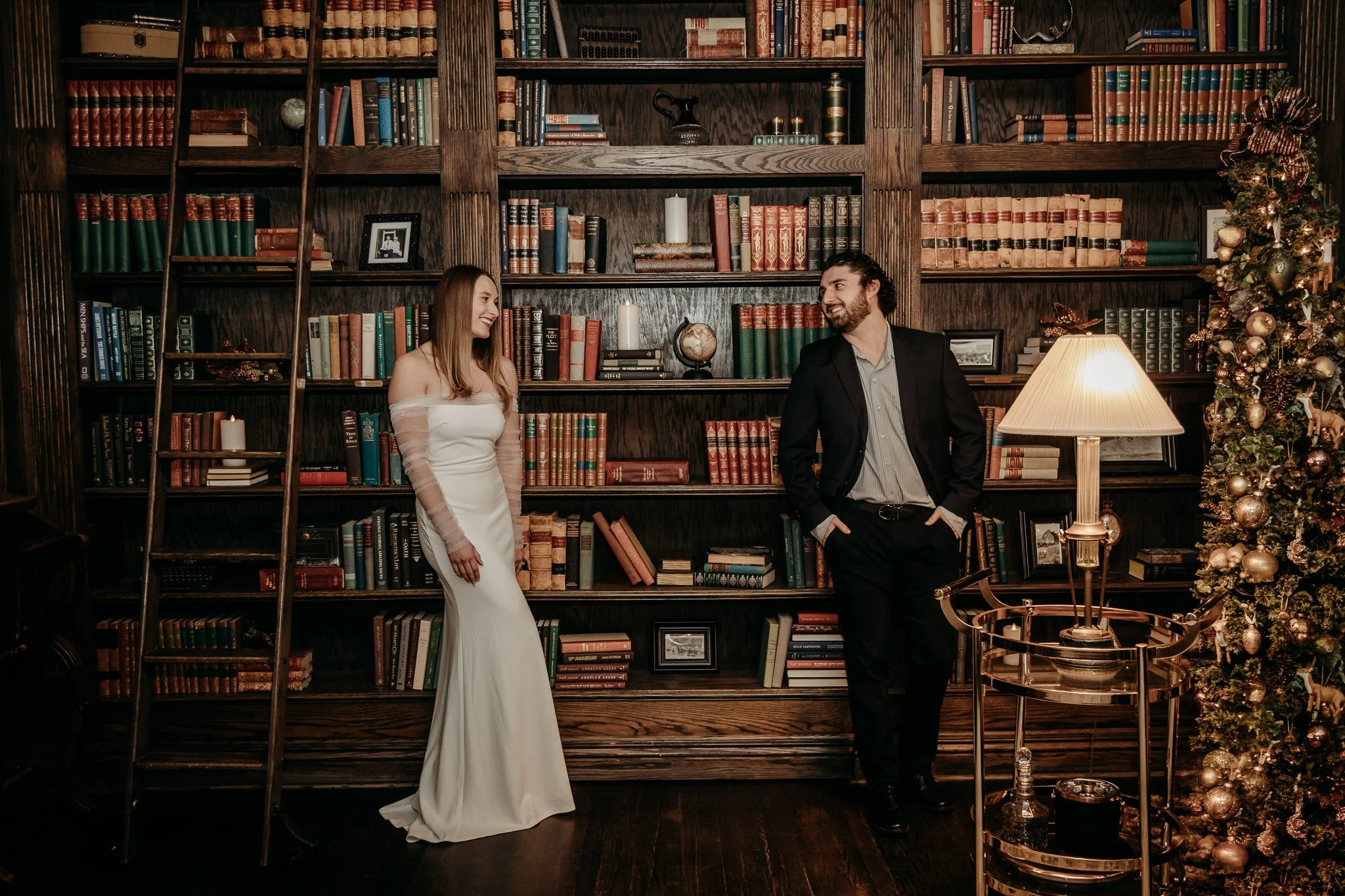 A man and a woman dressed in formal attire smiling and looking at each other inside a dimly lit room with a large wooden bookshelf filled with books. There is  a lamp on a side table.