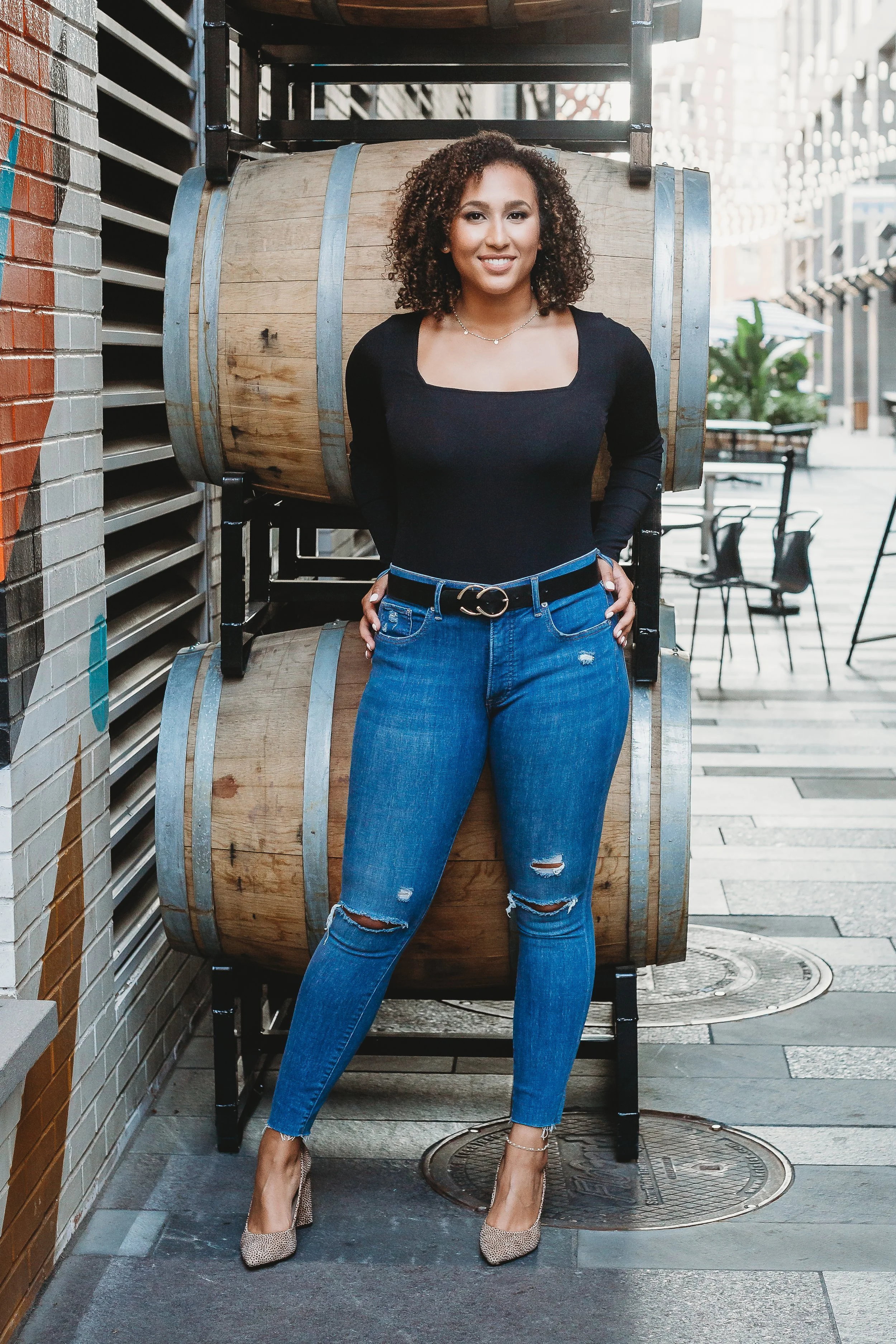 A woman with curly hair stands smiling in front of stacked large barrel drums on a city sidewalk, wearing a black long-sleeve top, distressed jeans, and high heels.