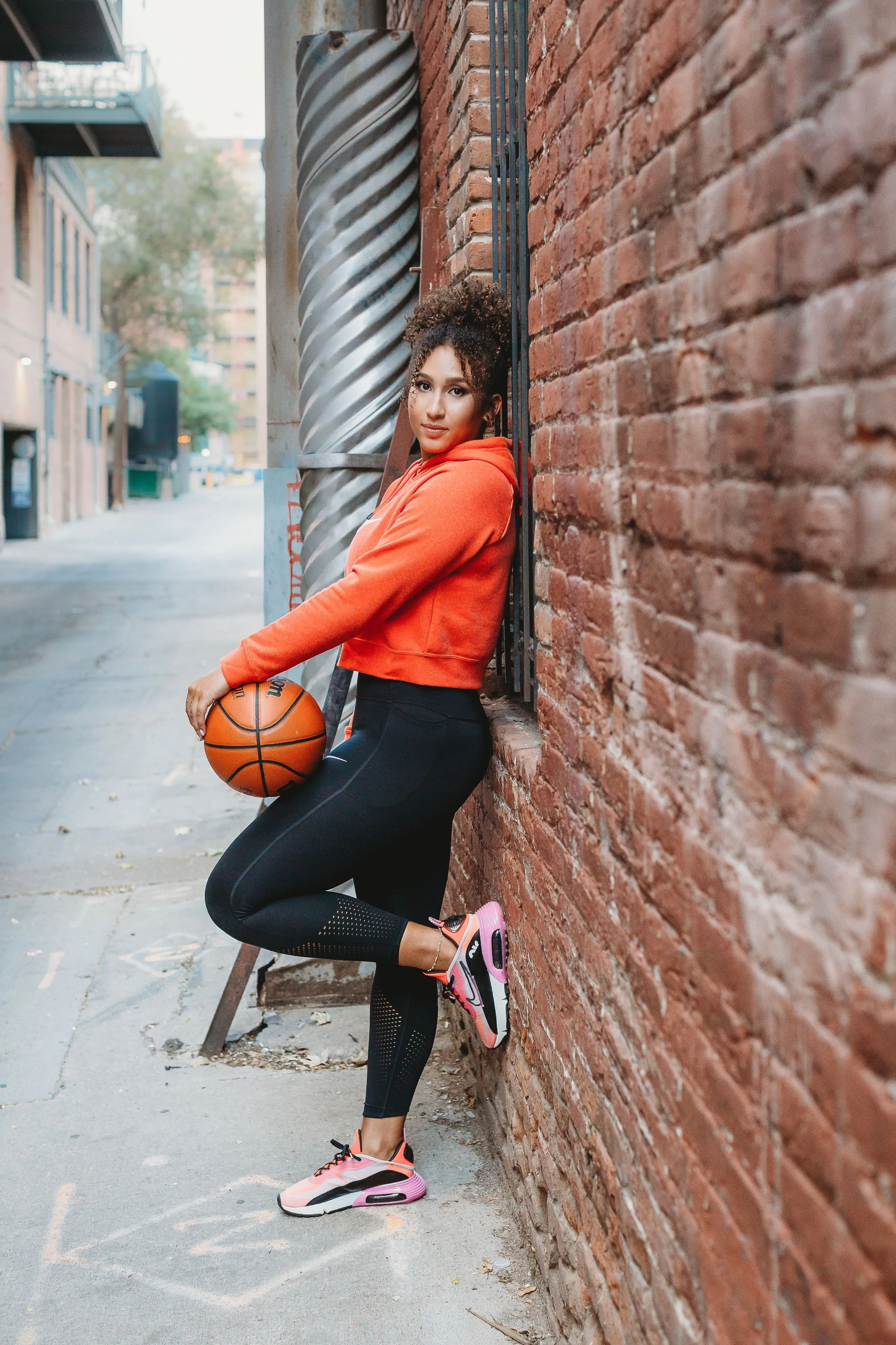 A young woman in athletic clothing, holding a basketball, leaning against a brick wall on an urban sidewalk.