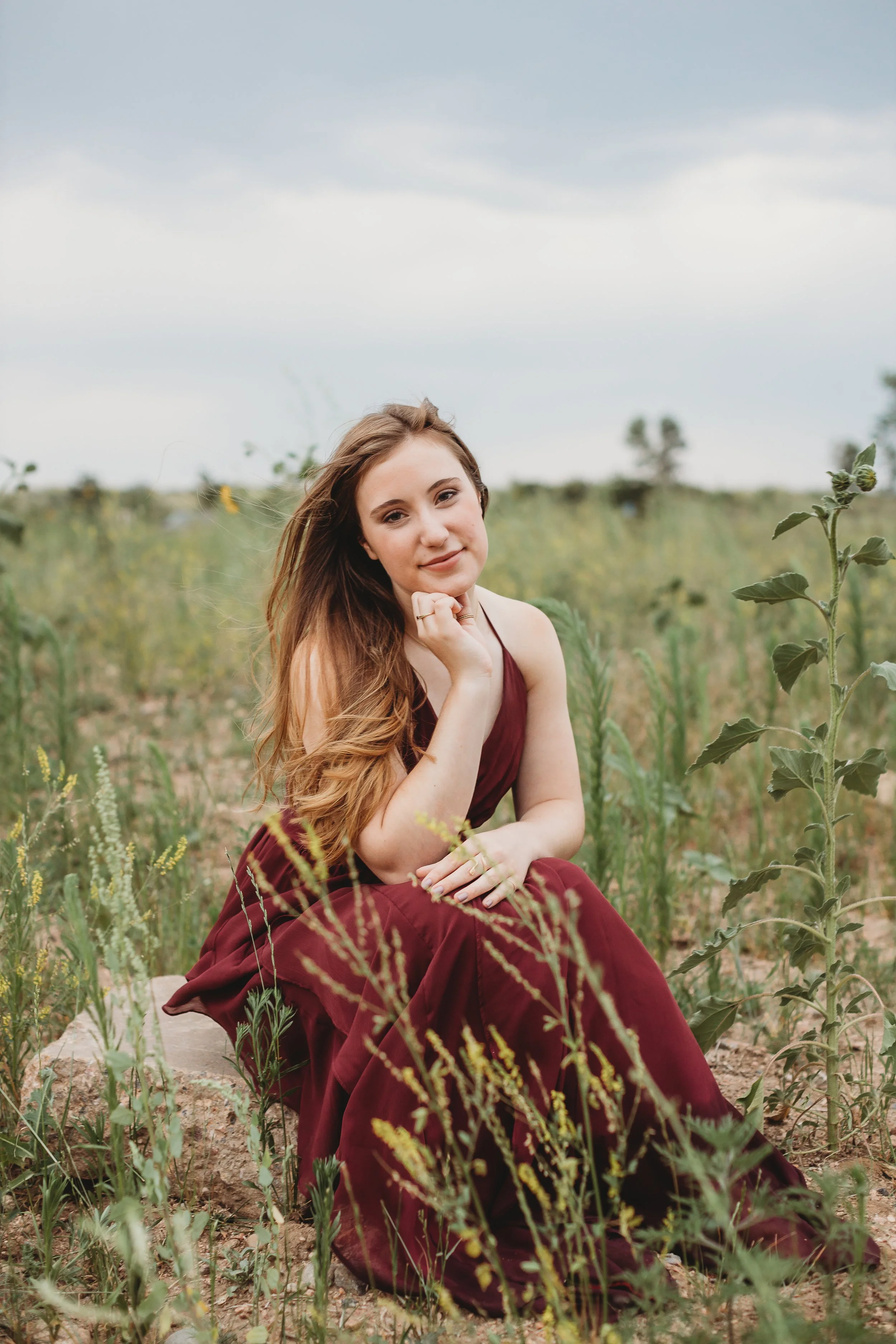 A young woman with long brown hair, wearing a maroon dress, is sitting on a rock in a field with green plants, during cloudy weather.