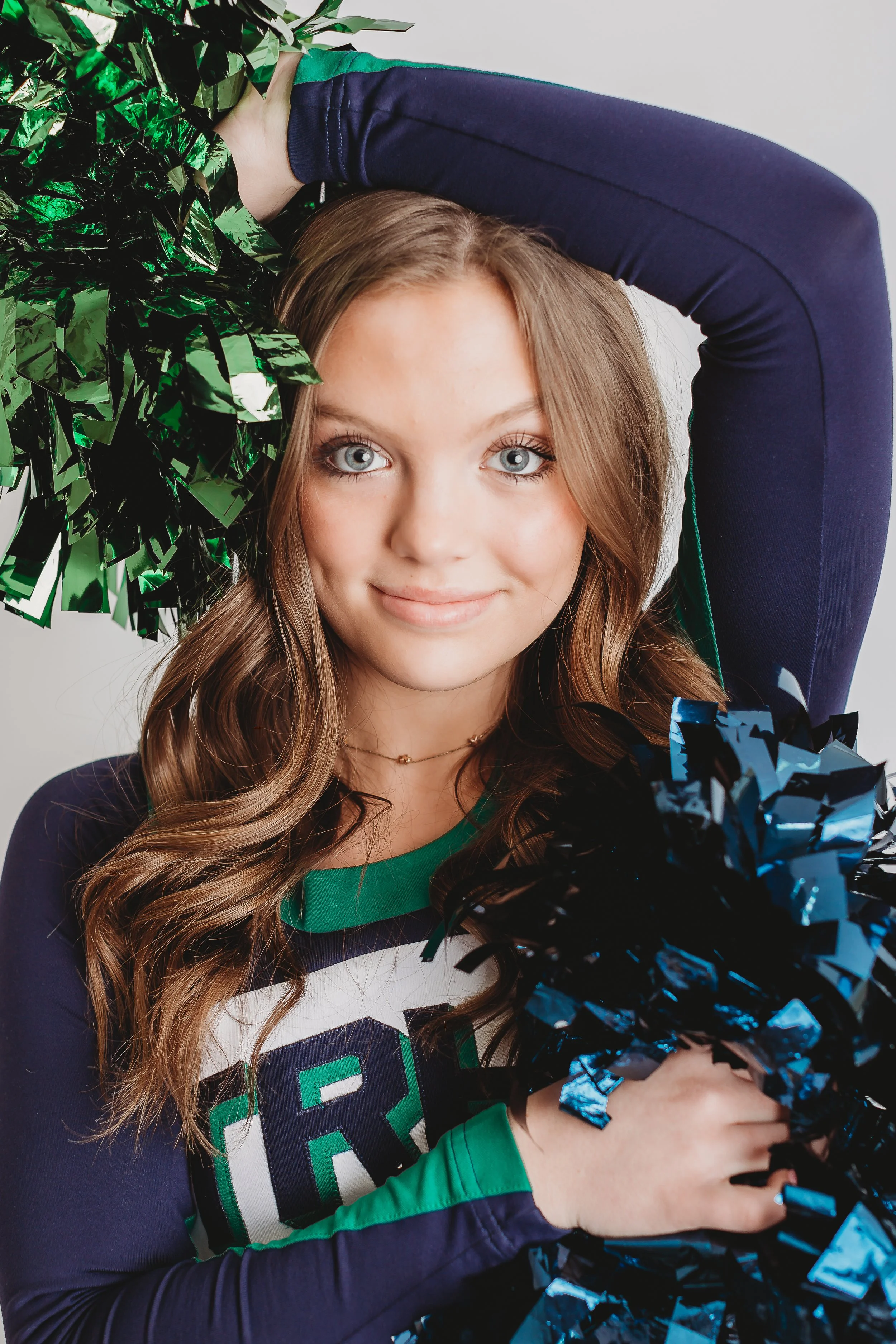 A young woman with long wavy brown hair and blue eyes smiling at the camera, holding blue and green metallic pom-poms, wearing a sports jersey with green, white, and blue colors.