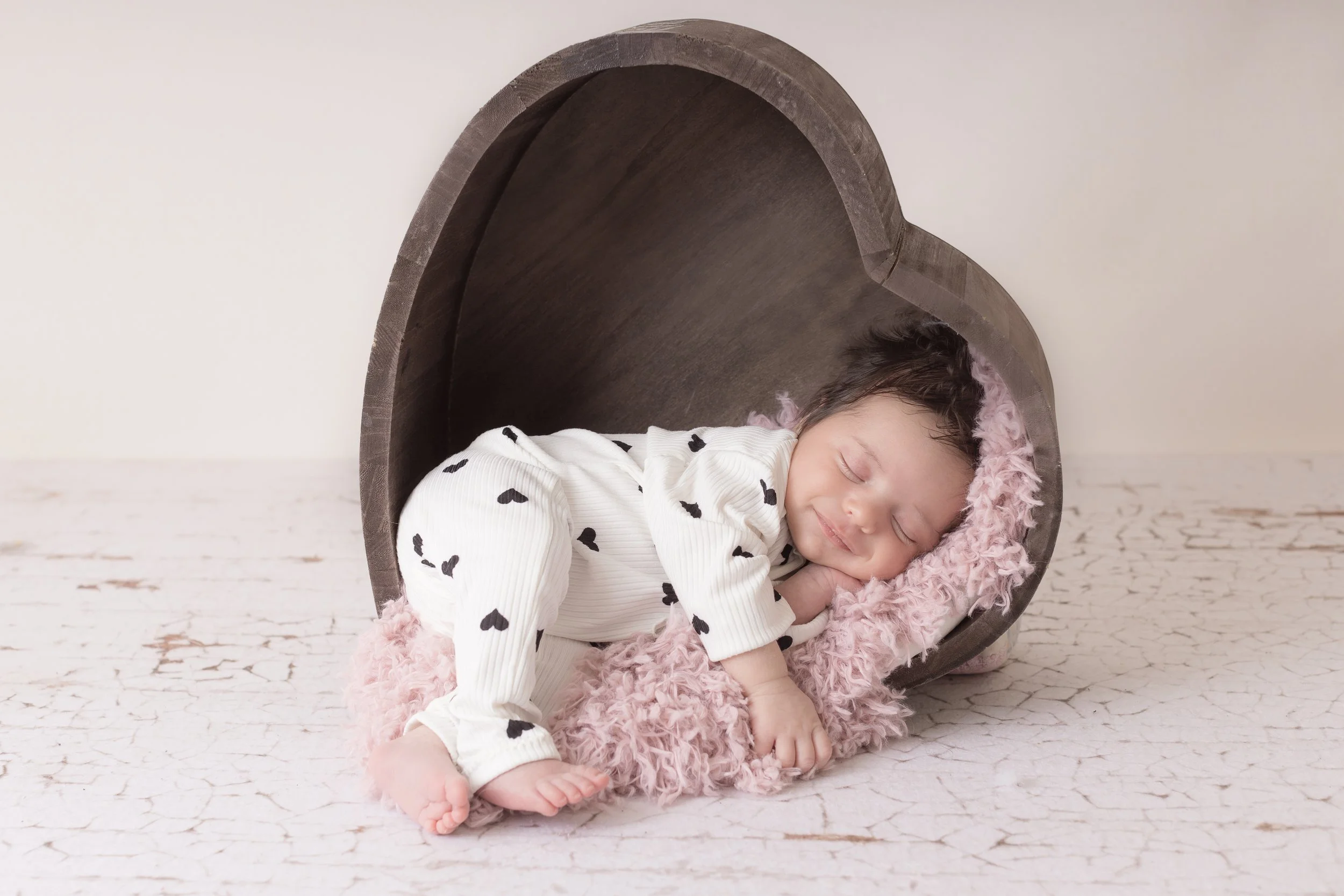 A sleeping baby girl with dark hair, wearing a white outfit with black heart patterns, lying on a soft pink fuzzy blanket inside a hollow wooden heart-shaped bed, on a distressed white floor.
