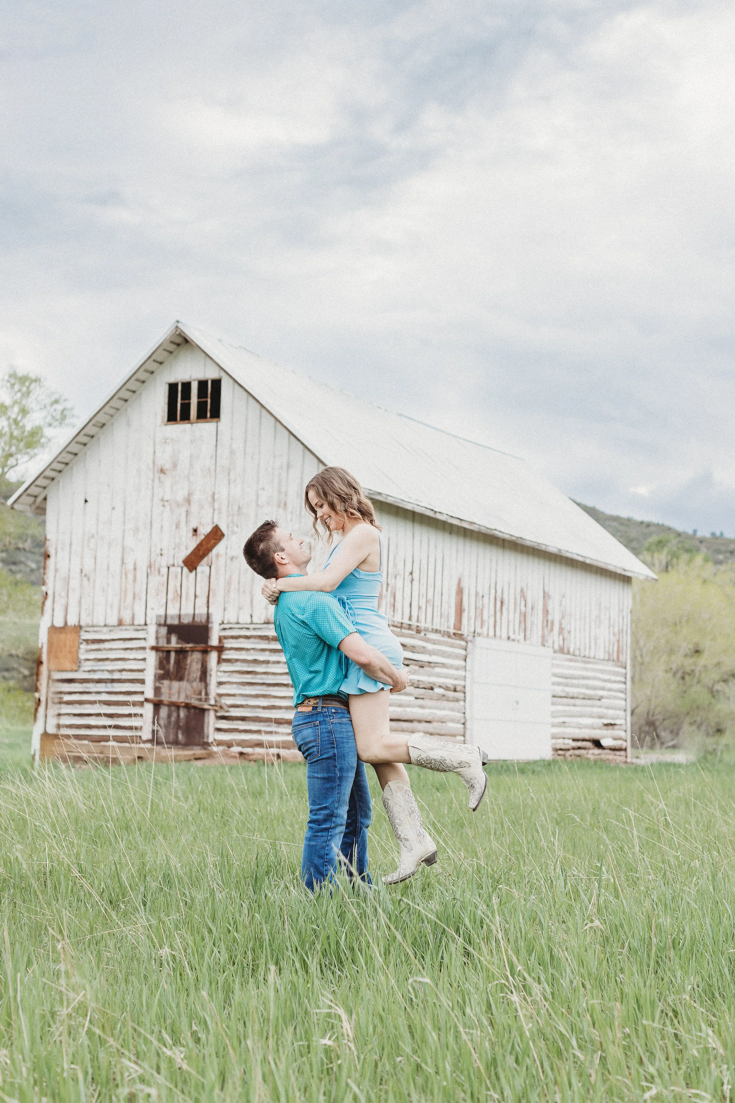 A couple is outside in a grassy field, with the man lifting the woman in his arms. The woman is wearing a light blue dress and white cowboy boots, and they are smiling at each other. An old, weathered barn is in the background.