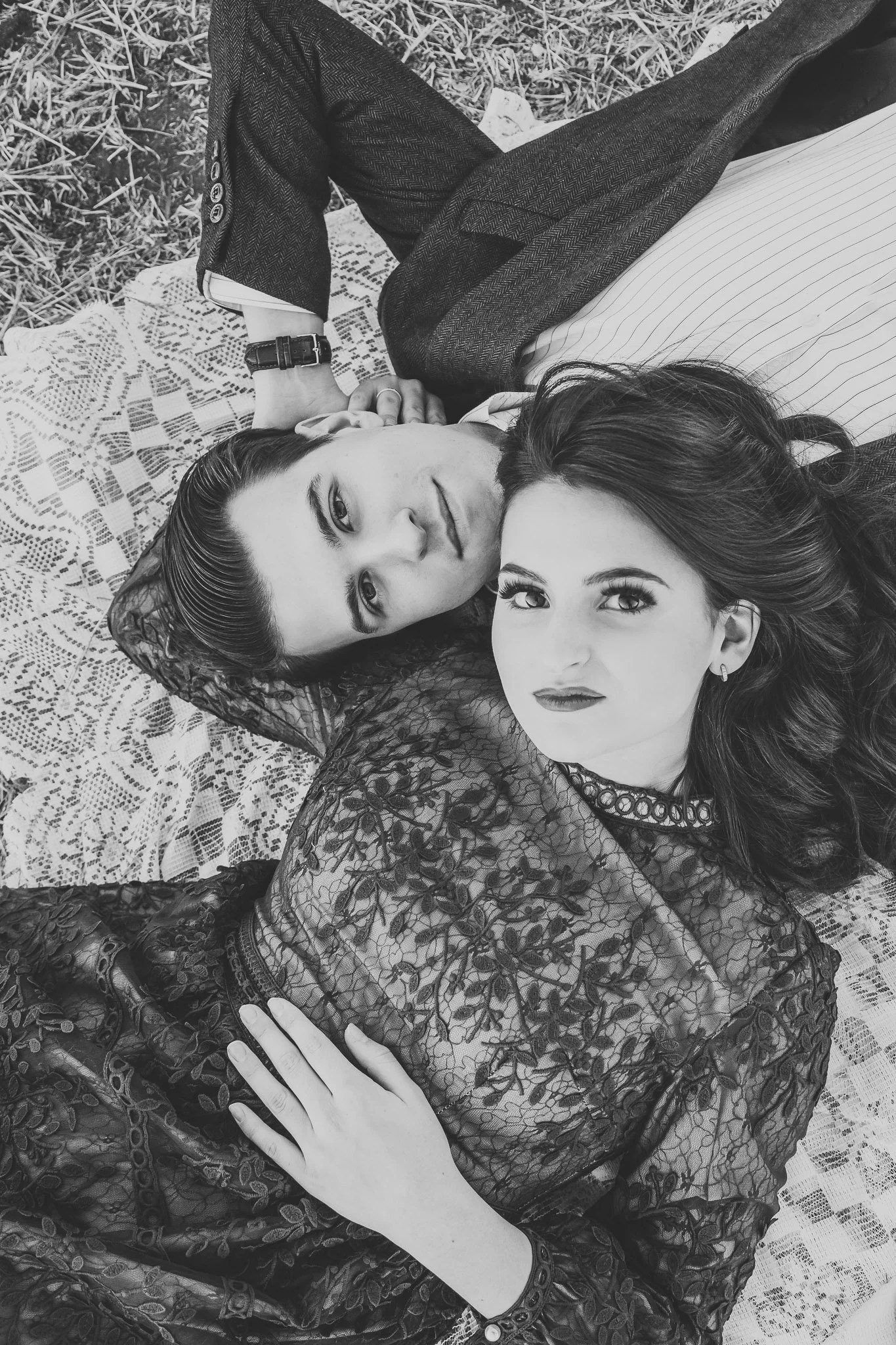 A black and white photo of a man and woman lying on a patterned blanket outdoors. The woman has long, wavy hair and is wearing a lacy dress, while the man has slicked-back hair and is dressed in a blazer over a striped shirt. They are looking at the camera, with the woman's hand resting on her lap.