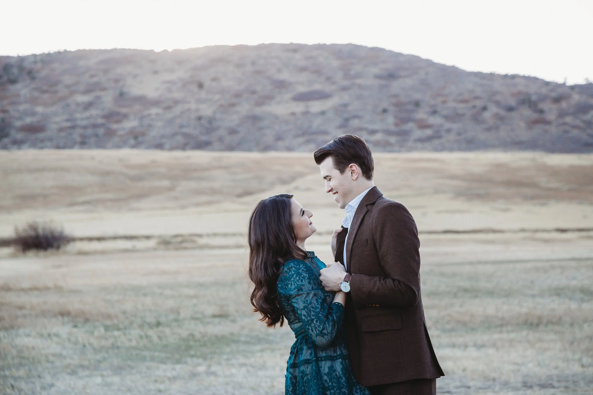 A man and woman standing close together outdoors, smiling at each other, with a grassy field and hills in the background.